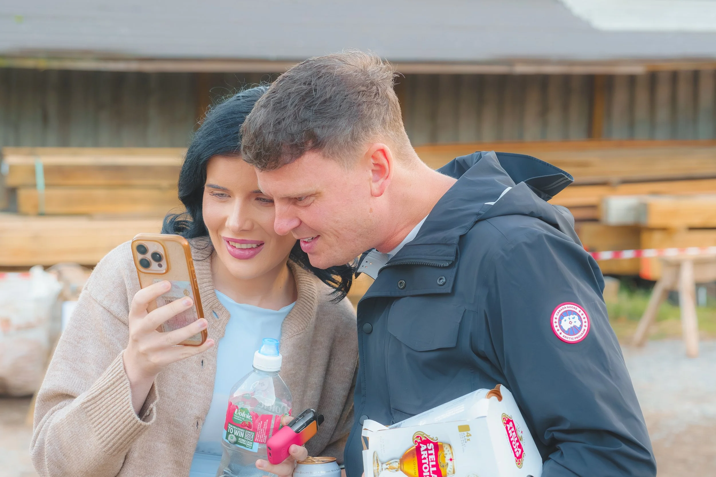A woman and a man are looking at a smartphone together, smiling. The woman is holding a bottle of flavored water, and the man is holding a bag of snack food. They appear to be outdoors, with wooden benches in the background, possibly at a market or e