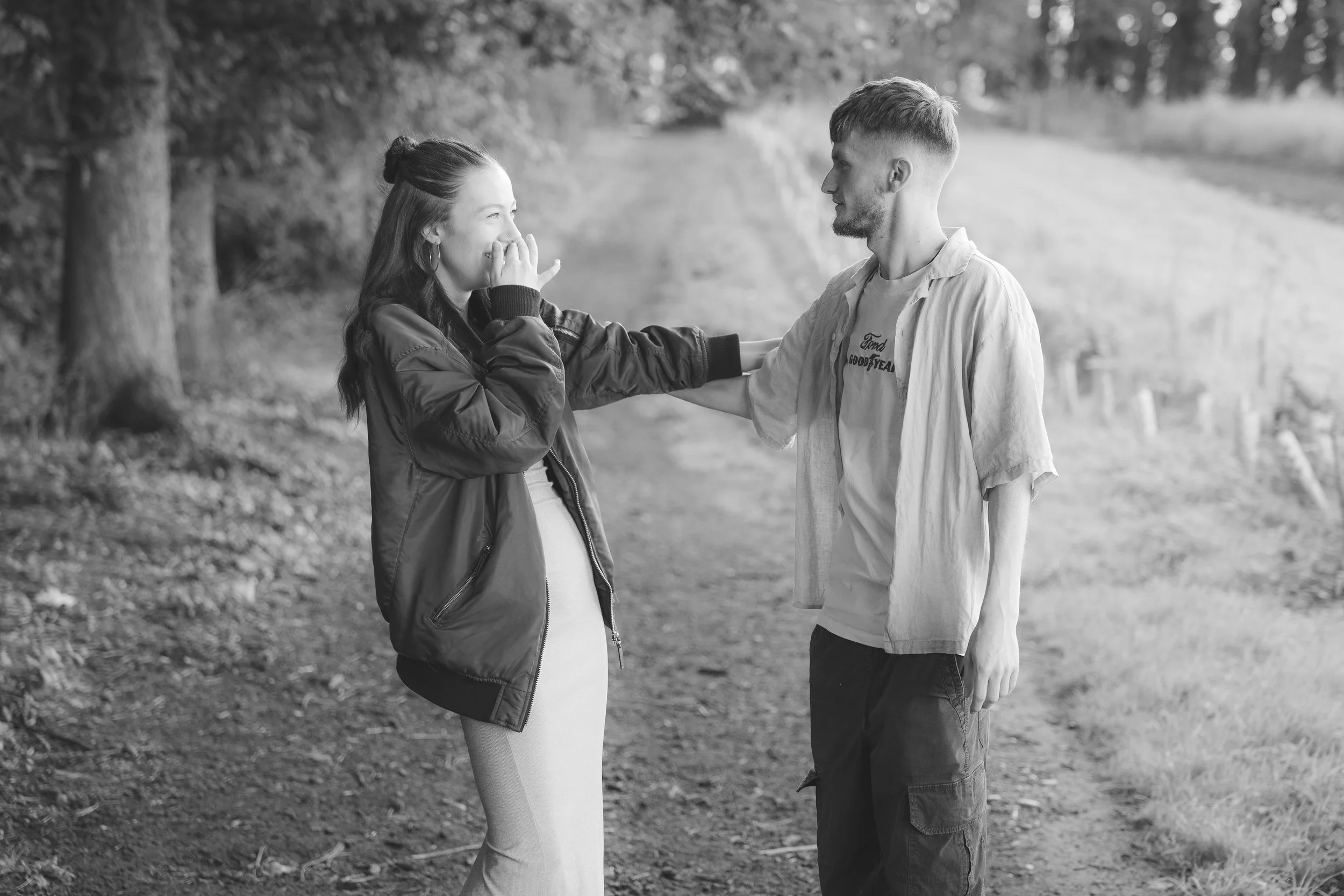 A black-and-white photo of a man and a pregnant woman standing outdoors near a lake, holding hands and looking at each other with smiles.