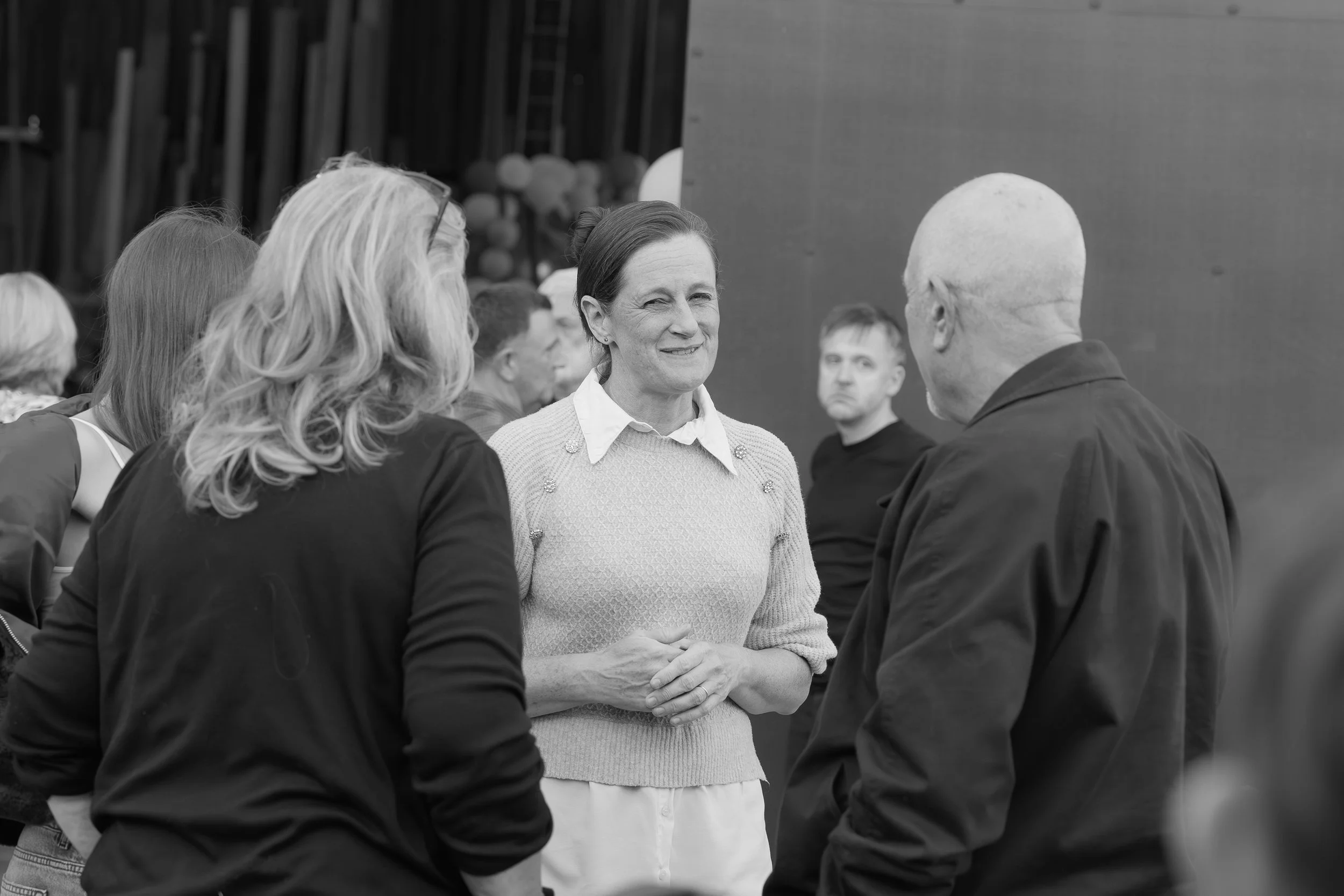 A woman with dark hair tied back, wearing a light-colored sweater, is engaged in a conversation with a man with short, light-colored hair, dressed in a dark jacket. Several other people are present in the background, suggesting a social or profession