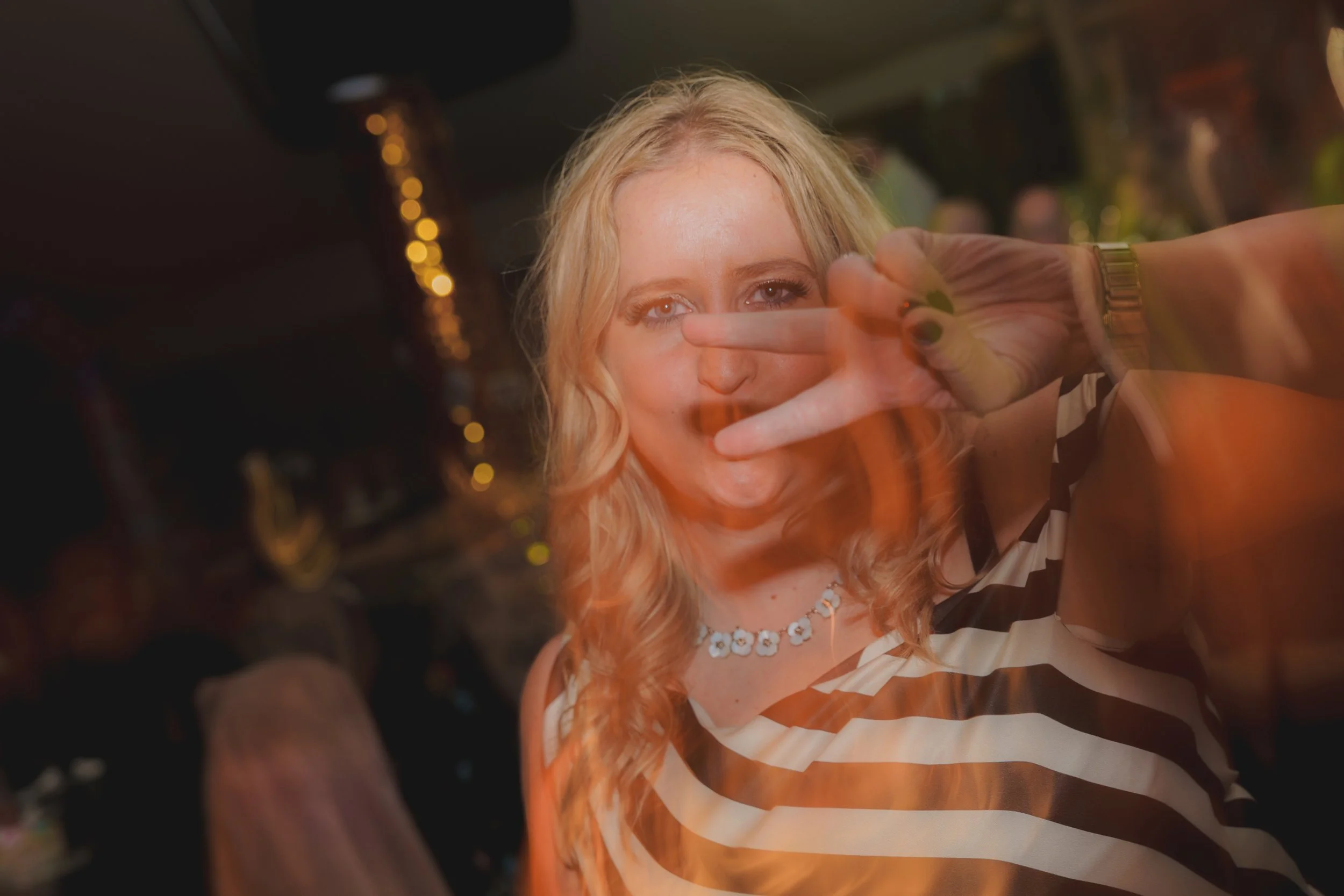 A woman with blonde curly hair making a peace sign with her fingers in front of her face, smiling towards the camera, with warm lighting and blurred background.