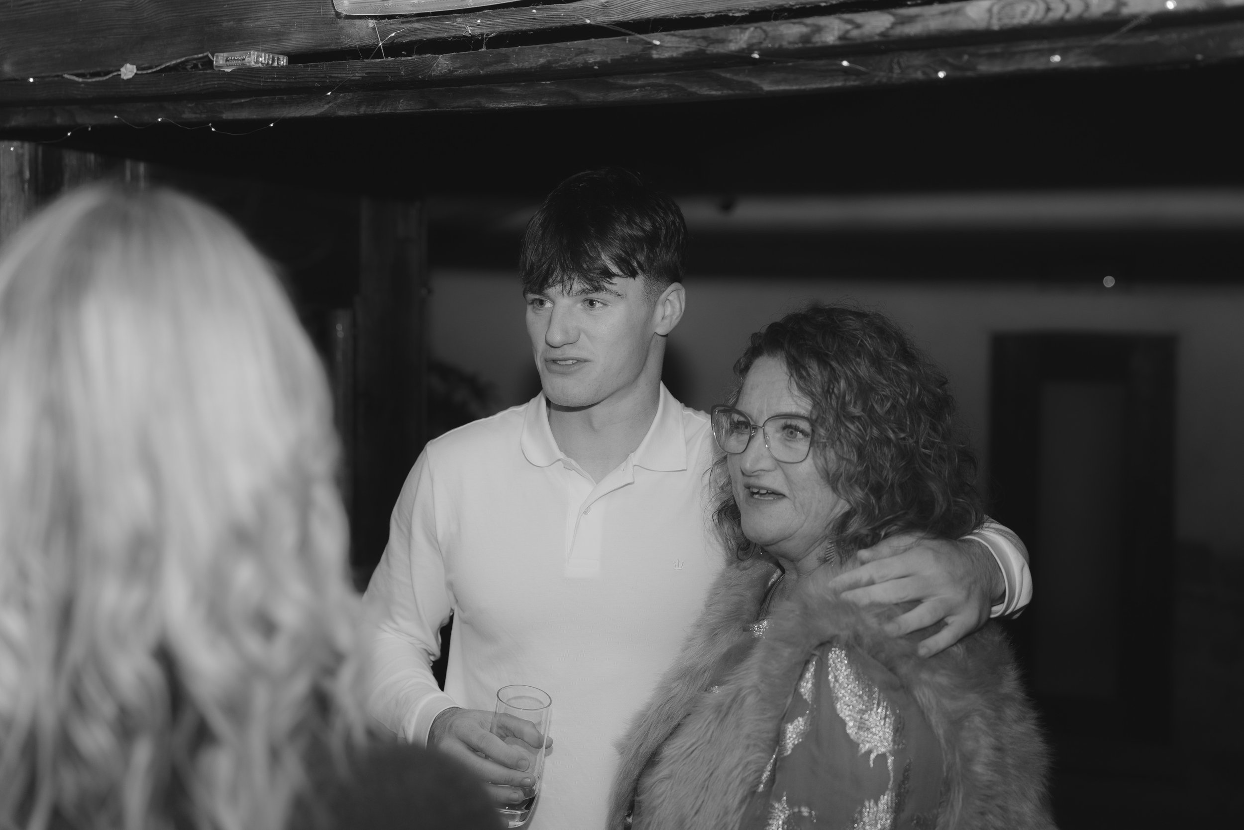 Three people are engaging in conversation at a social gathering. One man is holding a glass of drink and the other two women are wearing fur or faux fur coats. The image is in black and white.