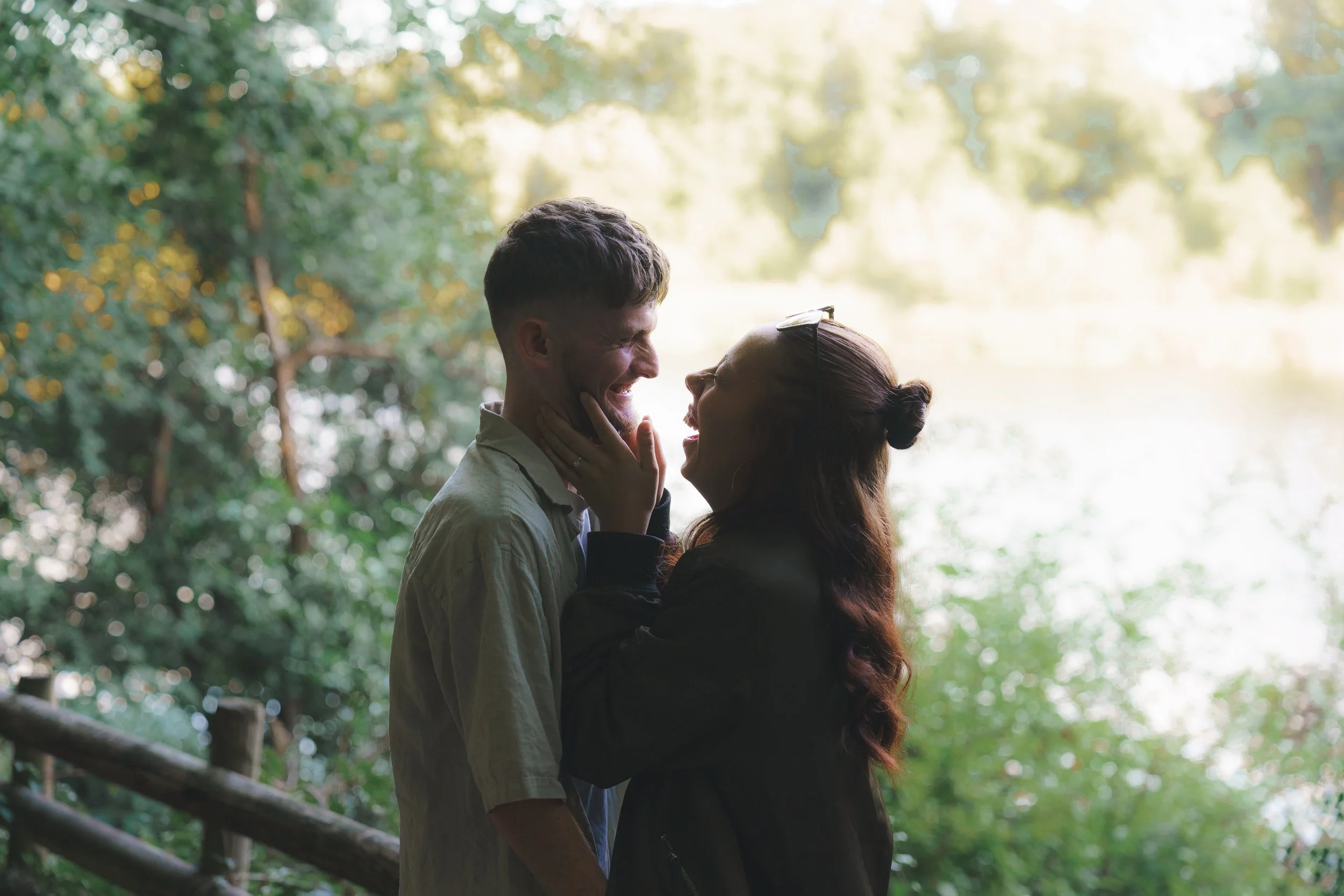 A couple standing outdoors near a lake, sharing a joyful moment and laughing together. The man is touching the woman's face, and she has her hands on his shoulders. It is a bright, sunny day with green trees in the background.