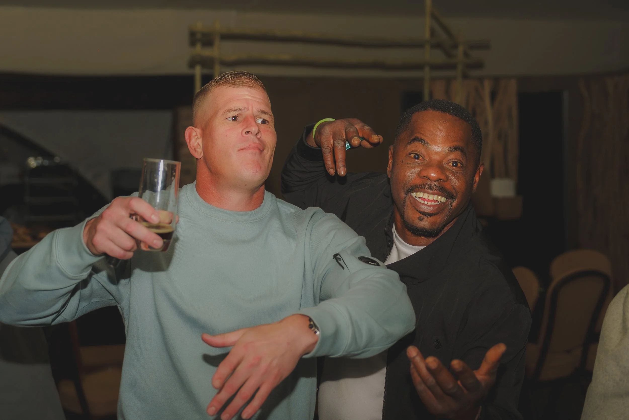 Two men are taking a selfie together, one holding a glass and making a peace sign with his fingers. They are smiling and appear to be enjoying themselves in a lively indoor setting.