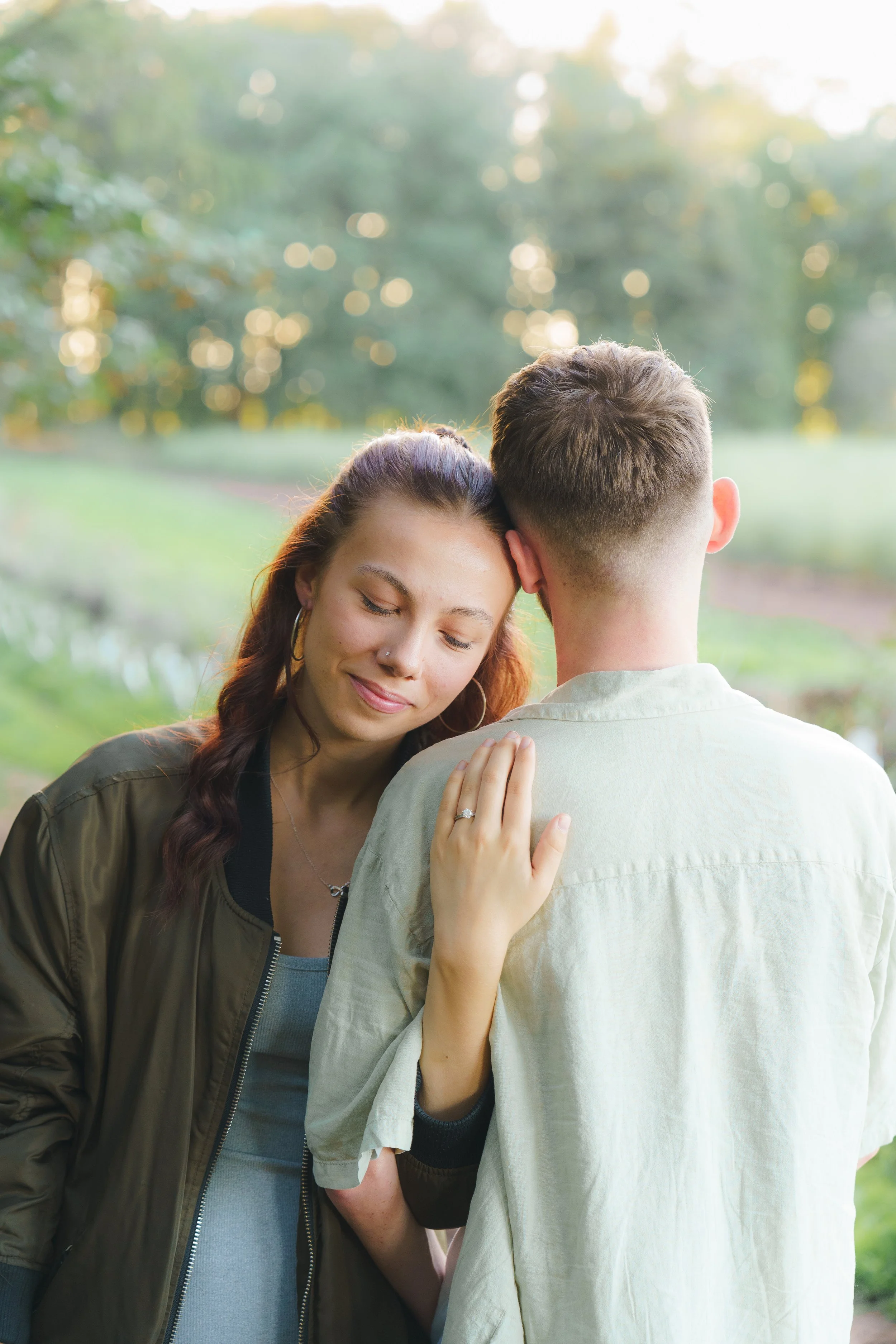 A young woman with long reddish-brown hair and hoop earrings is resting her head on the shoulder of a man with short light brown hair. She has her eyes closed and a gentle smile, with her hand on the man's shoulder, showing her engagement ring. They 