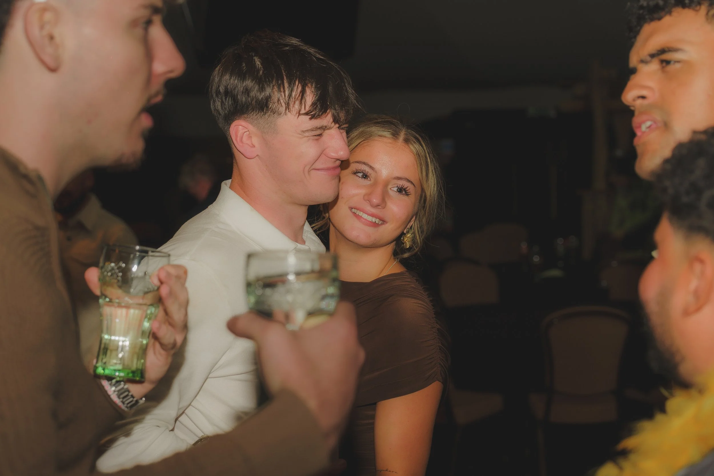 A group of young people in a social setting, with one man holding a glass of beer and smiling, while others are engaged in conversation, against a dark background.
