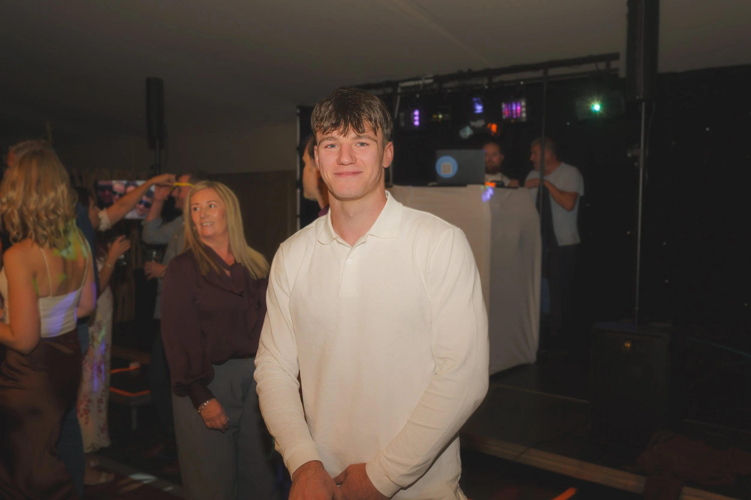 A young man in a white shirt smiling at a party or event, with people and DJ equipment in the background.