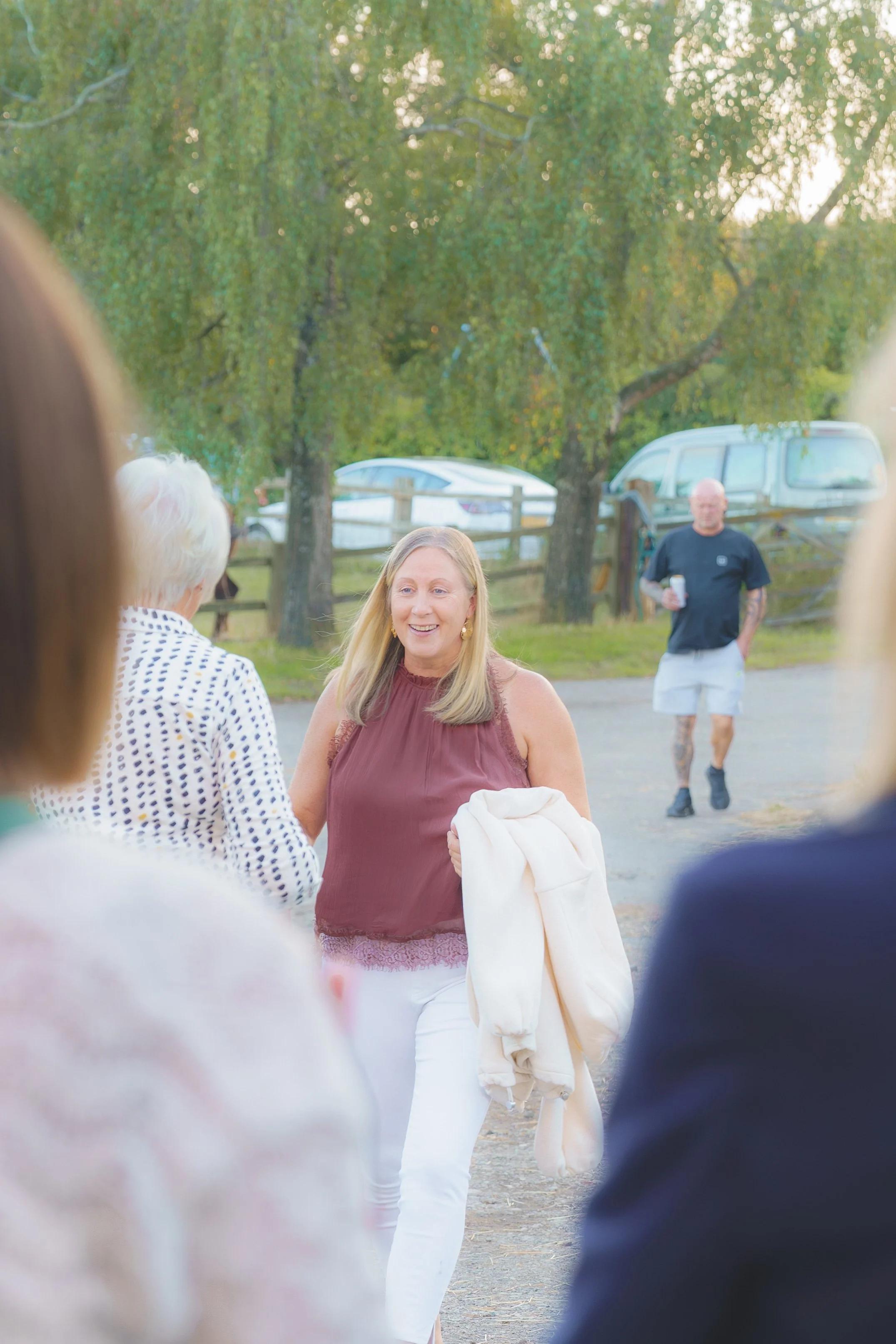 A woman with long blonde hair smiling and holding a white jacket, talking to an older woman with short white hair, who is wearing a white and black polka dot shirt, outdoors in a park-like setting with trees and parked cars in the background. A bald 