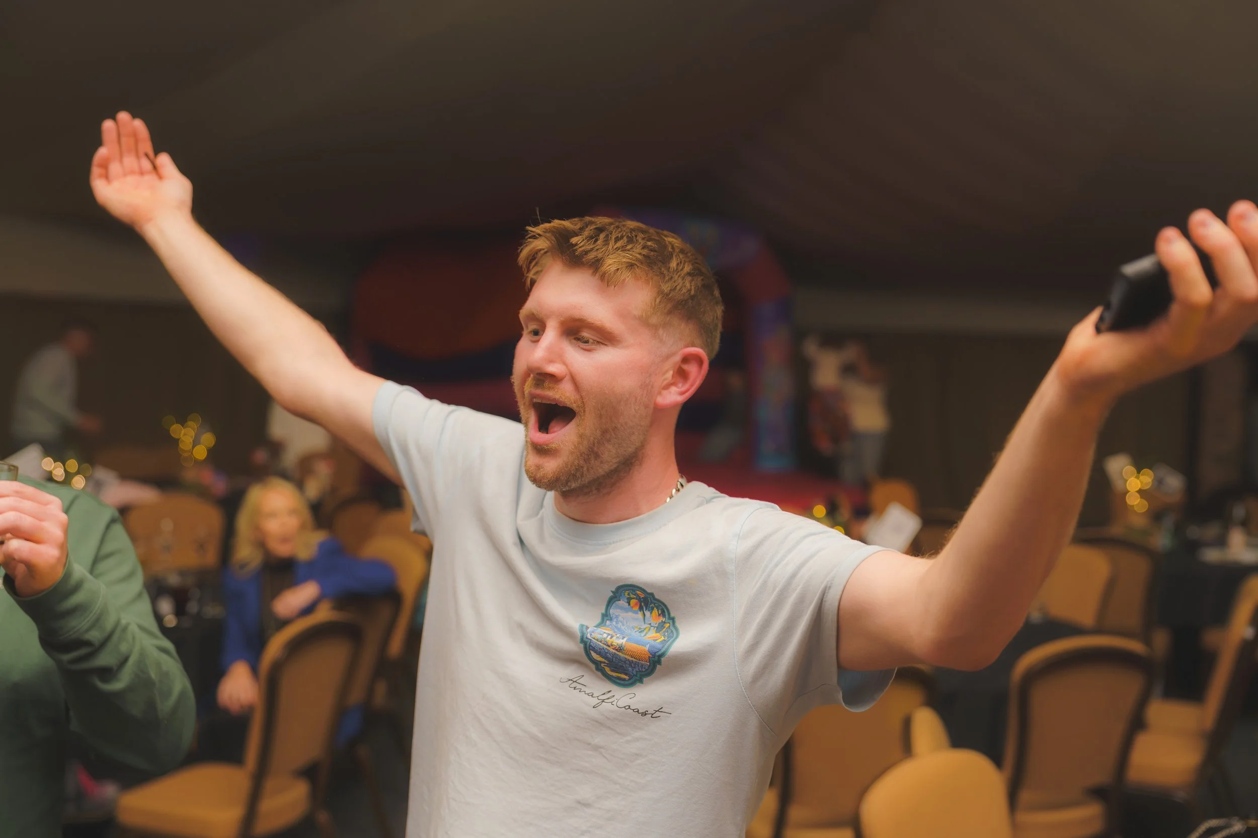 A young man with short reddish hair and a beard, wearing a white t-shirt with a colorful patch, is dancing with his arms raised and head tilted back, in a lively indoor setting with people seated at tables.