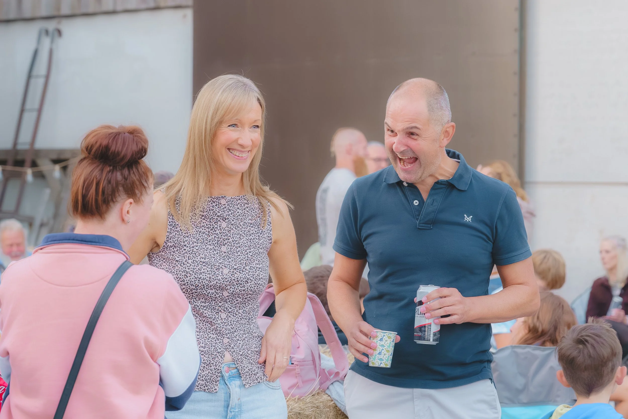Man and woman smiling and talking at an outdoor gathering, surrounded by children and other adults.
