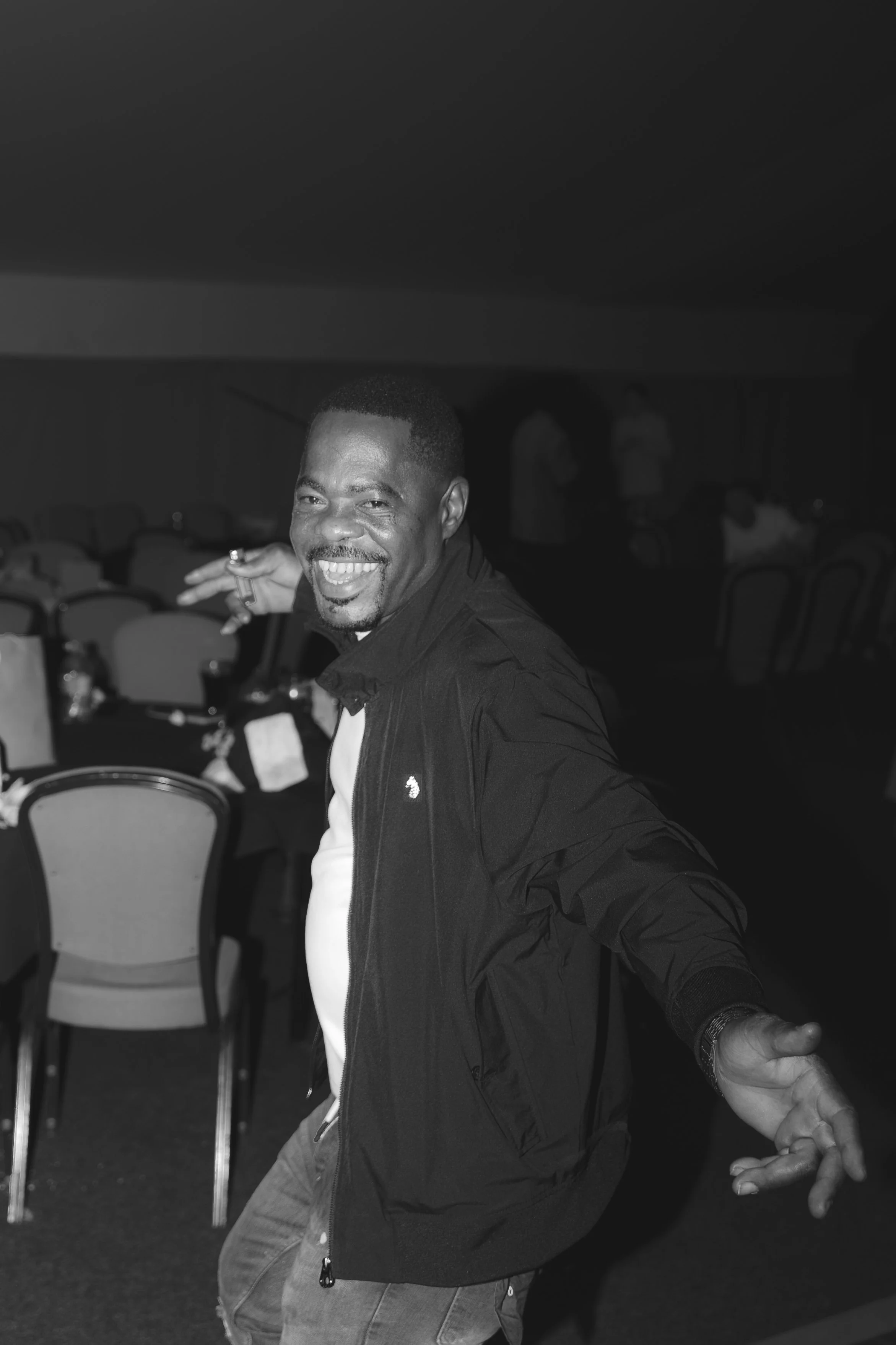 A man dancing and smiling at a social gathering, with chairs and other people in the background.