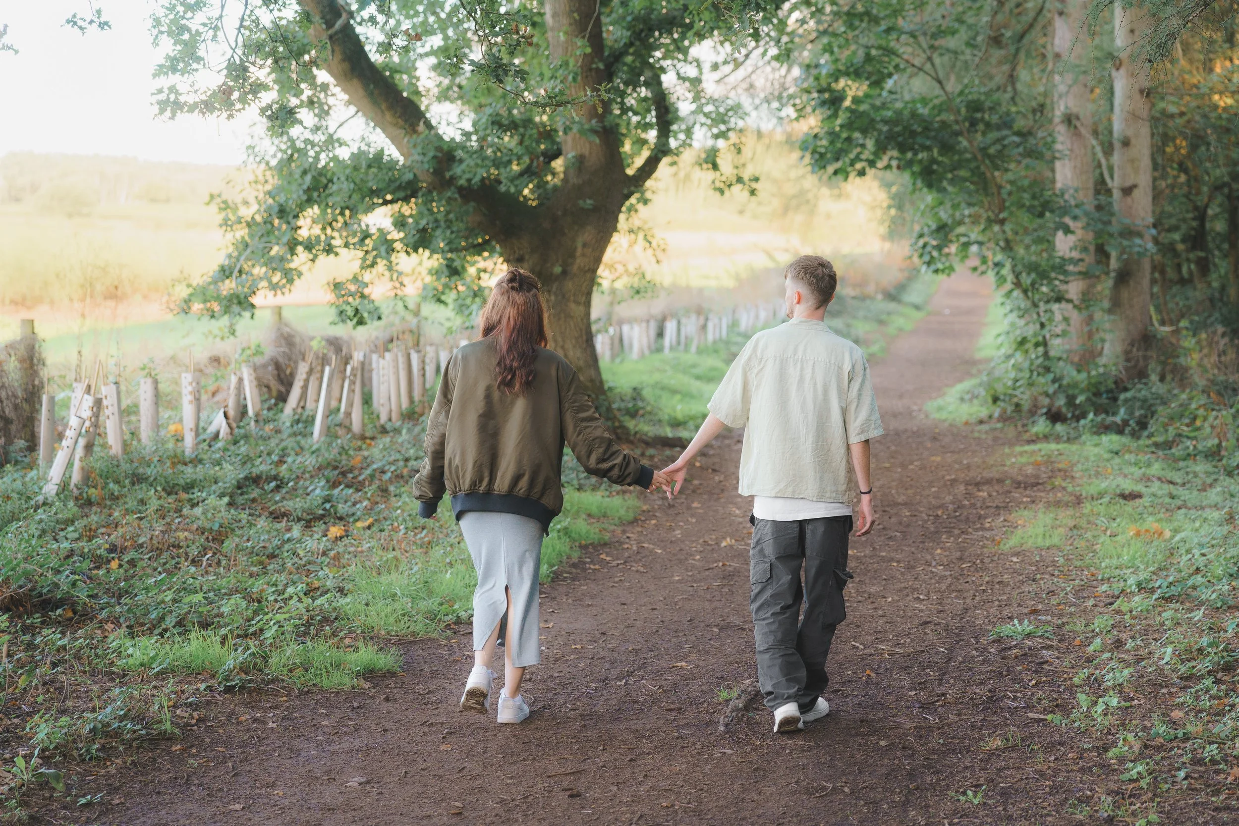 A couple walking hand in hand on a dirt path through a green wooded area during daytime.