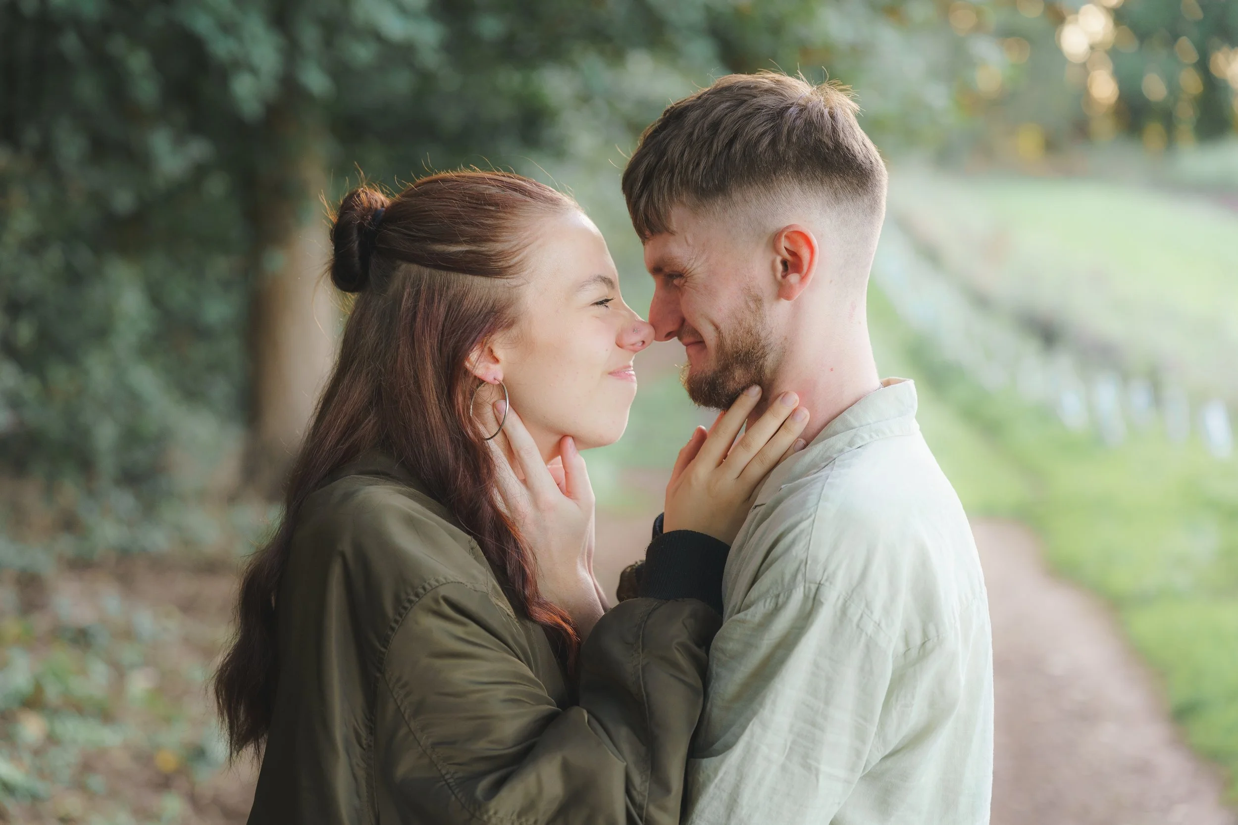 A young couple standing close to each other in a park, smiling with their foreheads touching and eyes closed, holding each other's faces affectionately.