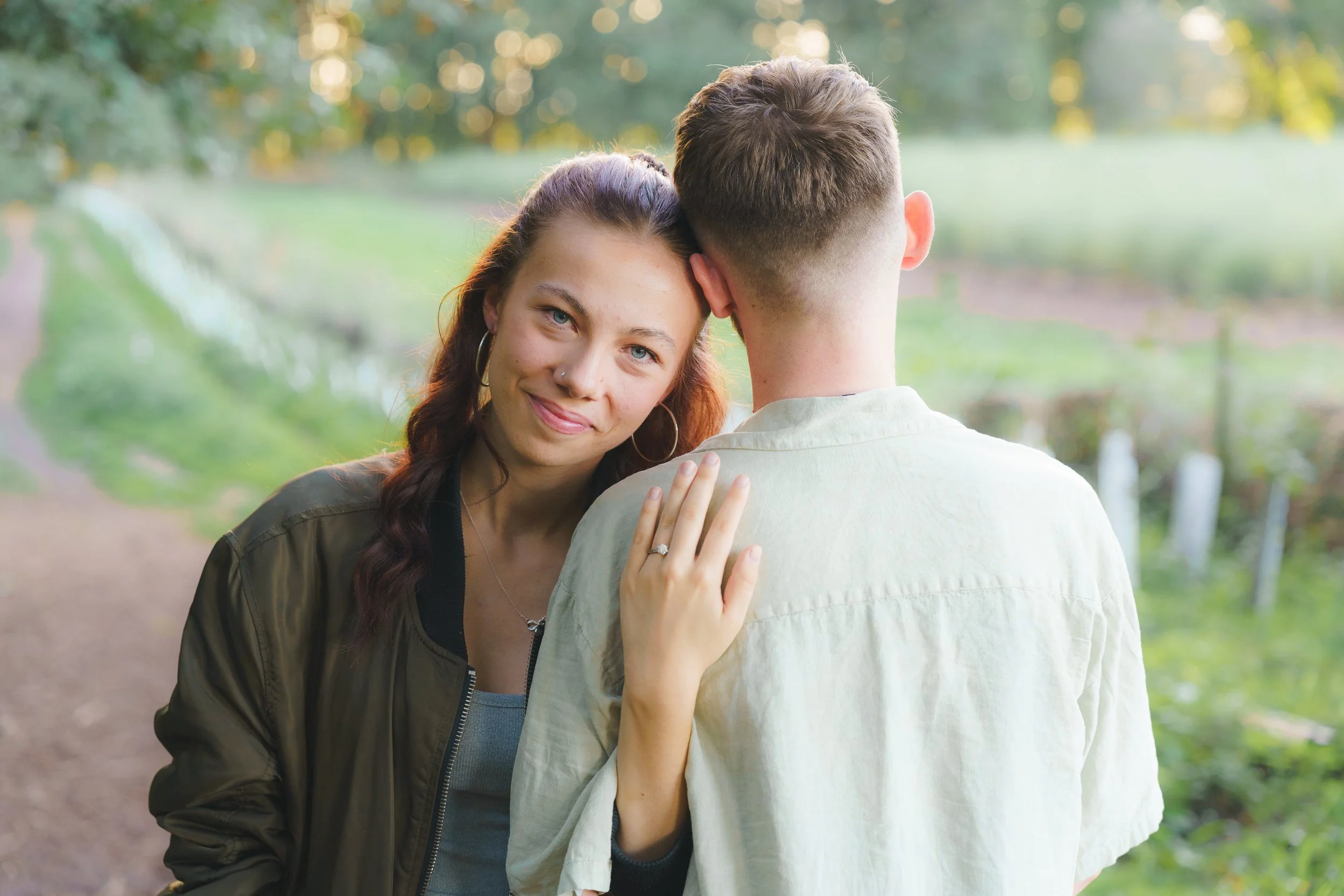A woman with long, dark hair, earrings, and a nose ring smiling and resting her head on a man’s shoulder with his back facing the camera, outdoors in a park with trees and a pathway.