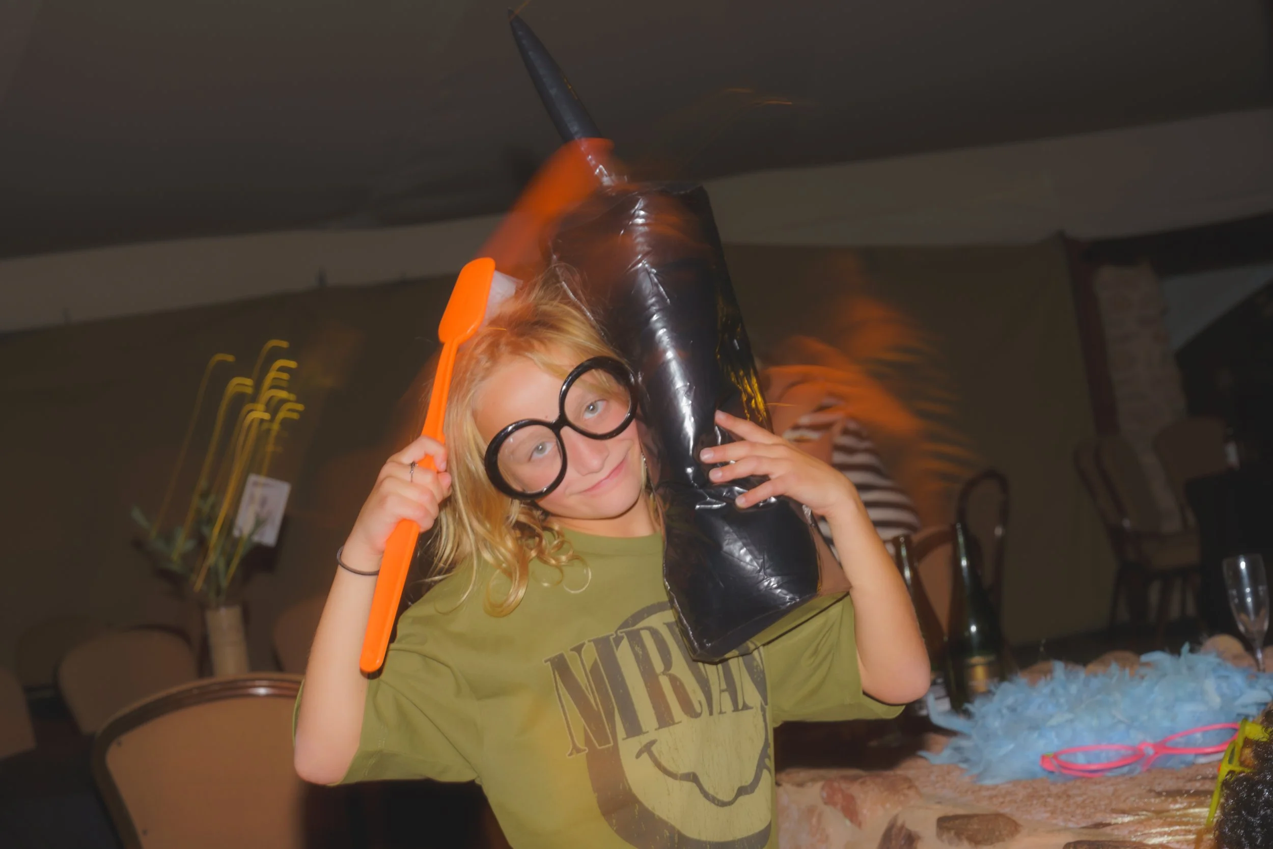 A young girl with blonde hair and glasses holding a toy light saber, standing in a room with a wrapped black gift and other party supplies on a table.