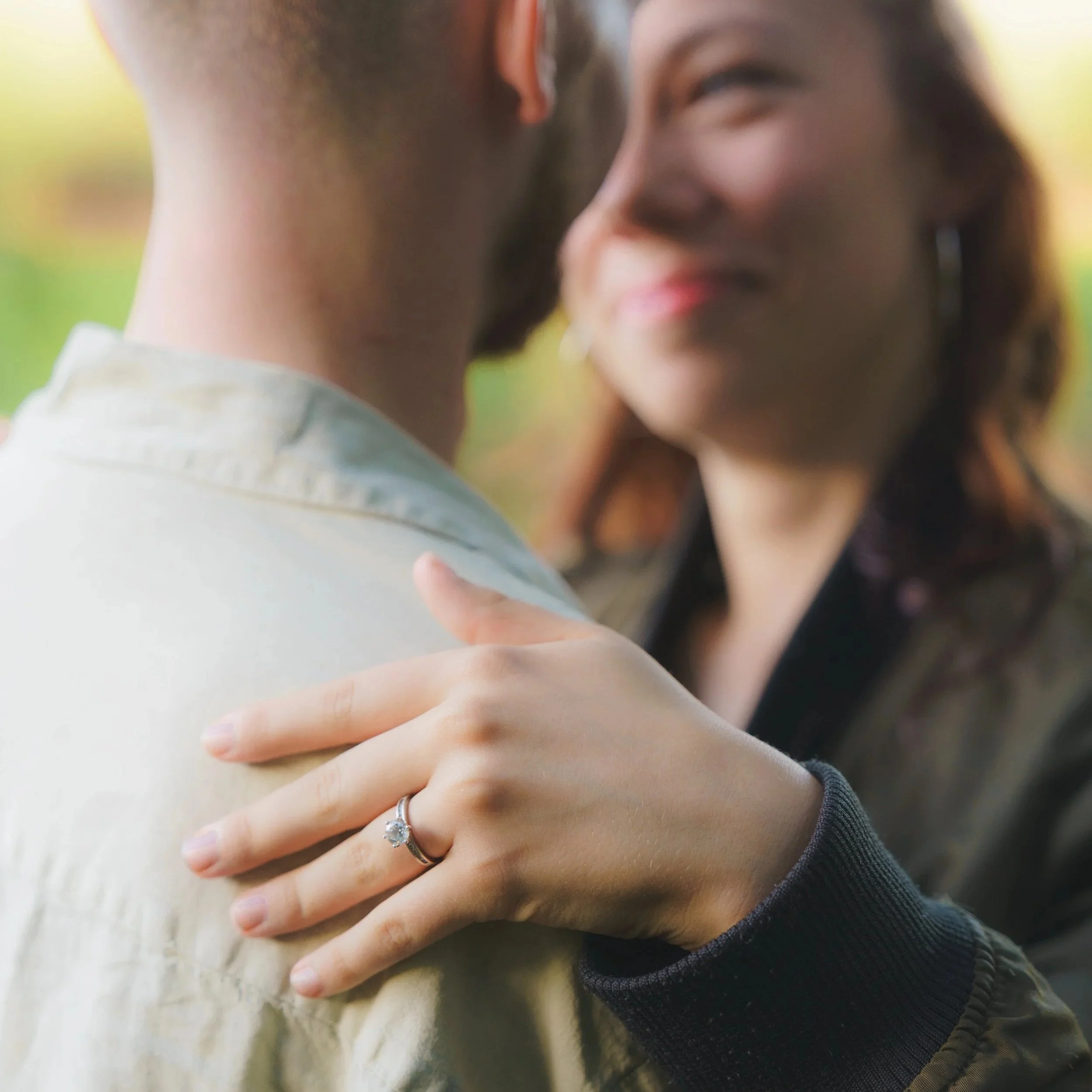 A close-up of a woman and man embracing, with the woman's hand showing an engagement ring. The woman has a smile and dark hair, and they are outside with a blurred natural background.