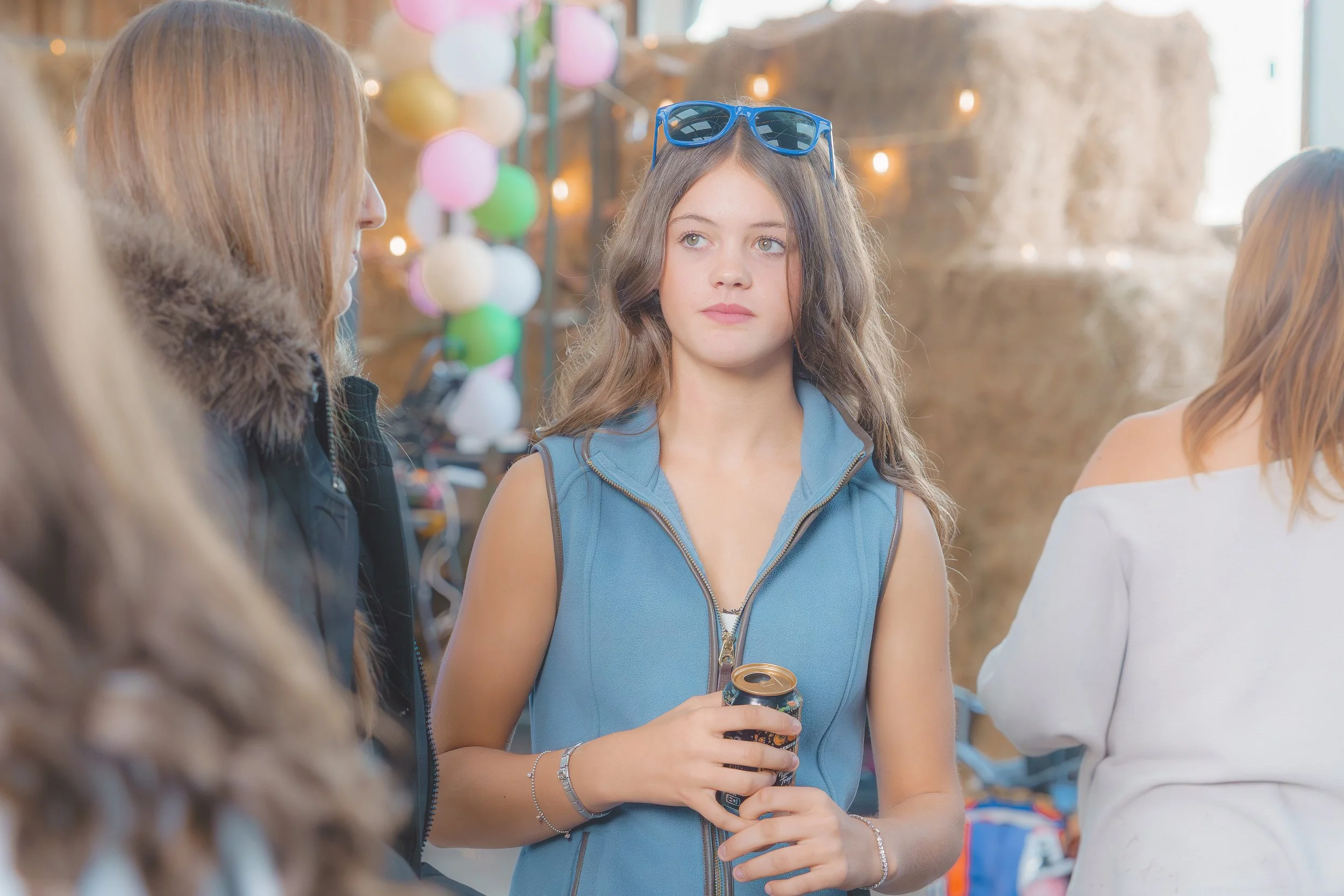 A young girl with wavy brown hair, glasses on her head, holding a soda can, standing in a decorated area with colorful balloons and string lights, engaging in conversation with other people.