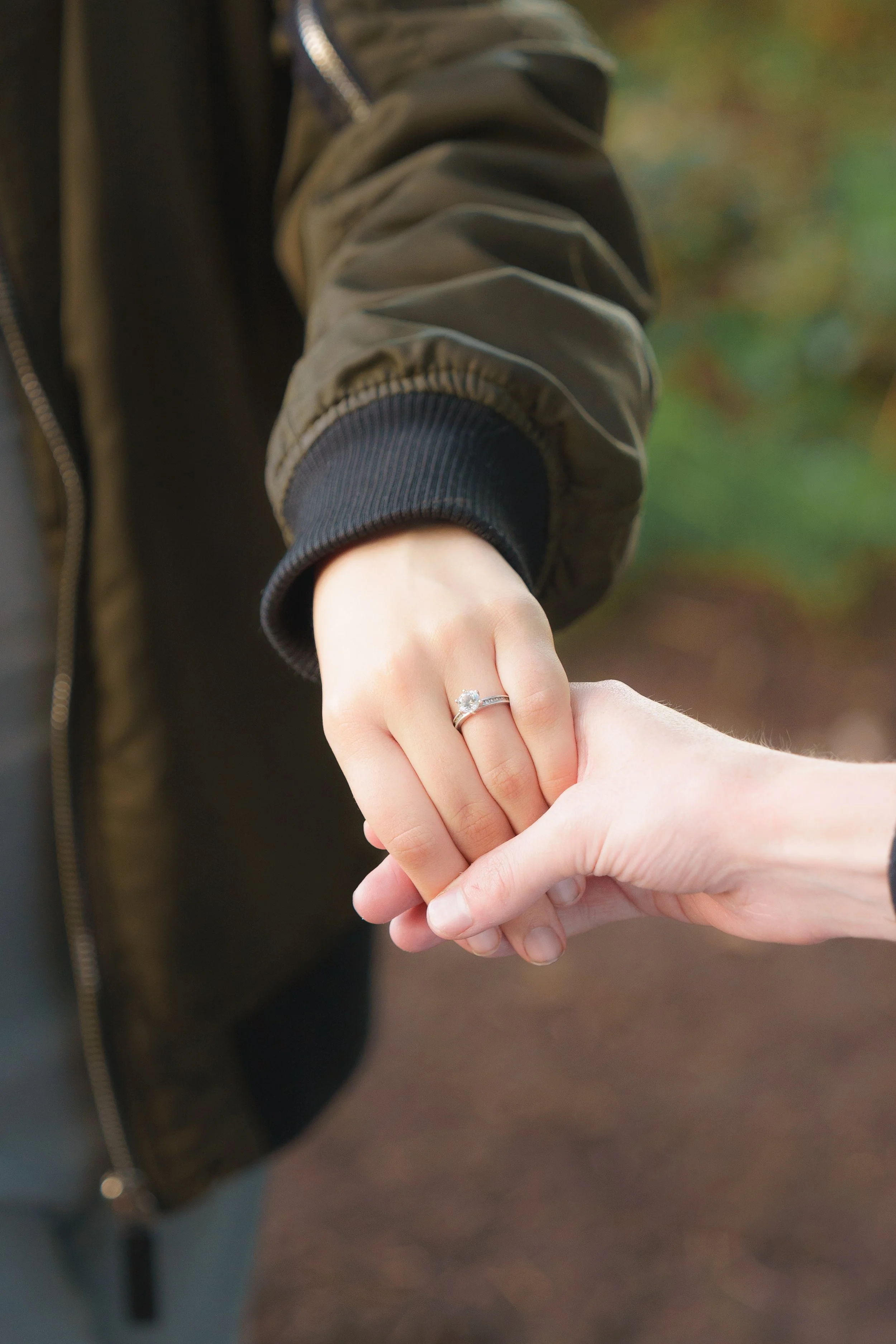 Close-up of two people holding hands, with one person wearing a ring with a large gem, and the other person's hand being gently grasped. The person with the ring is wearing a dark jacket with a striped cuff.