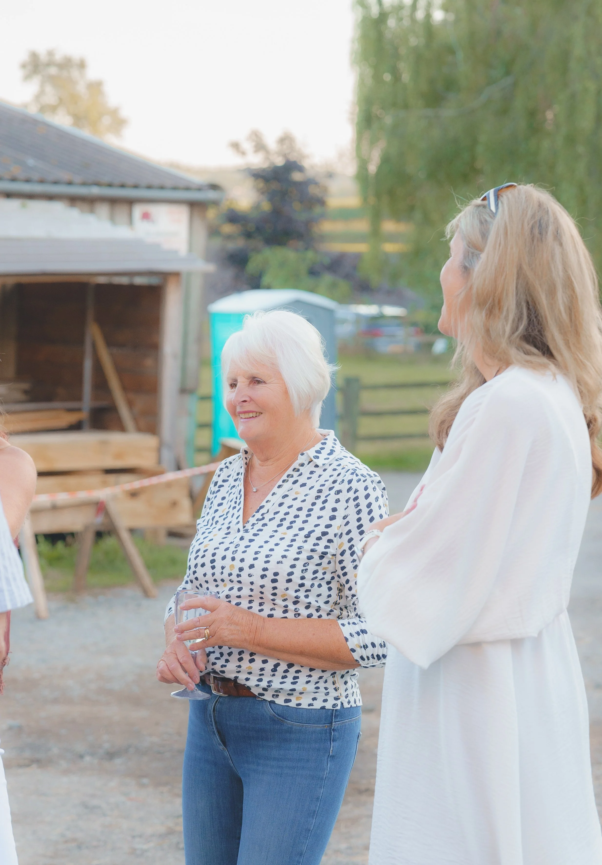 Two women having a conversation outdoors during daytime; one older woman with short white hair, wearing a polka dot shirt and jeans, holding a wine glass, and the other woman with long blonde hair, wearing a white dress, glasses on her head.