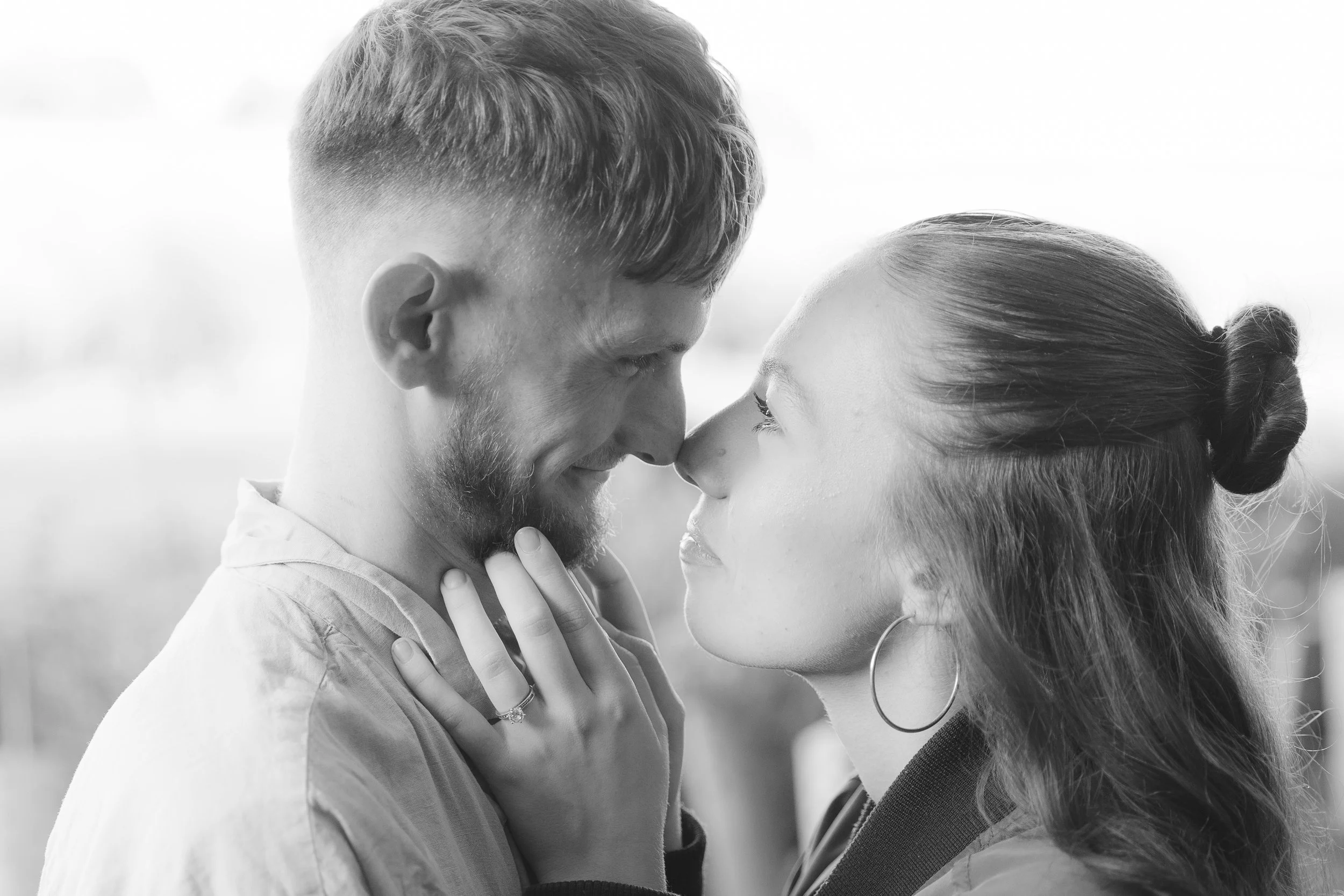 A black and white close-up photo of a couple facing each other with their foreheads touching and eyes closed. The man has short hair and a beard, and the woman has long hair pulled back. They appear to be sharing an intimate moment.