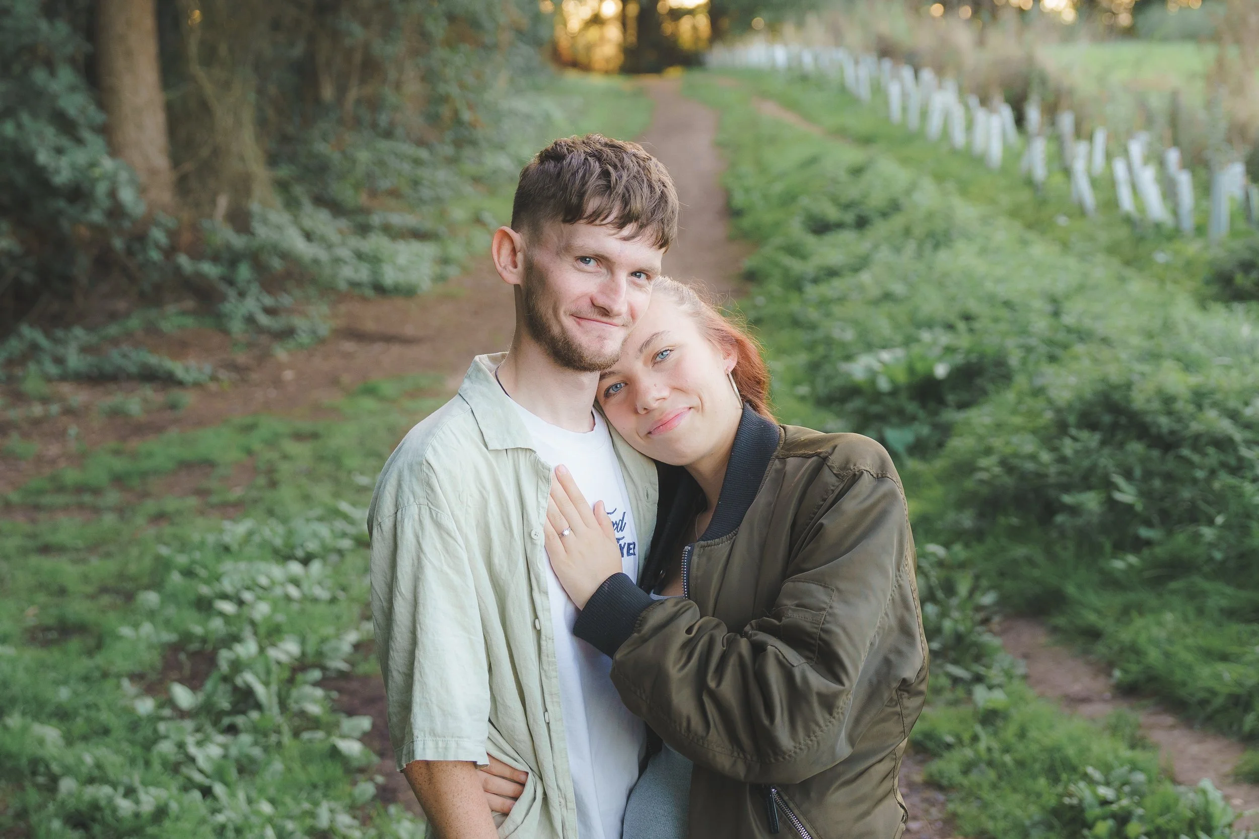 A young couple embracing on a trail in a green, wooded area during daytime. The man has short brown hair and wears a light-colored jacket, while the woman has reddish hair and wears a dark green jacket. They are both smiling and appear happy.