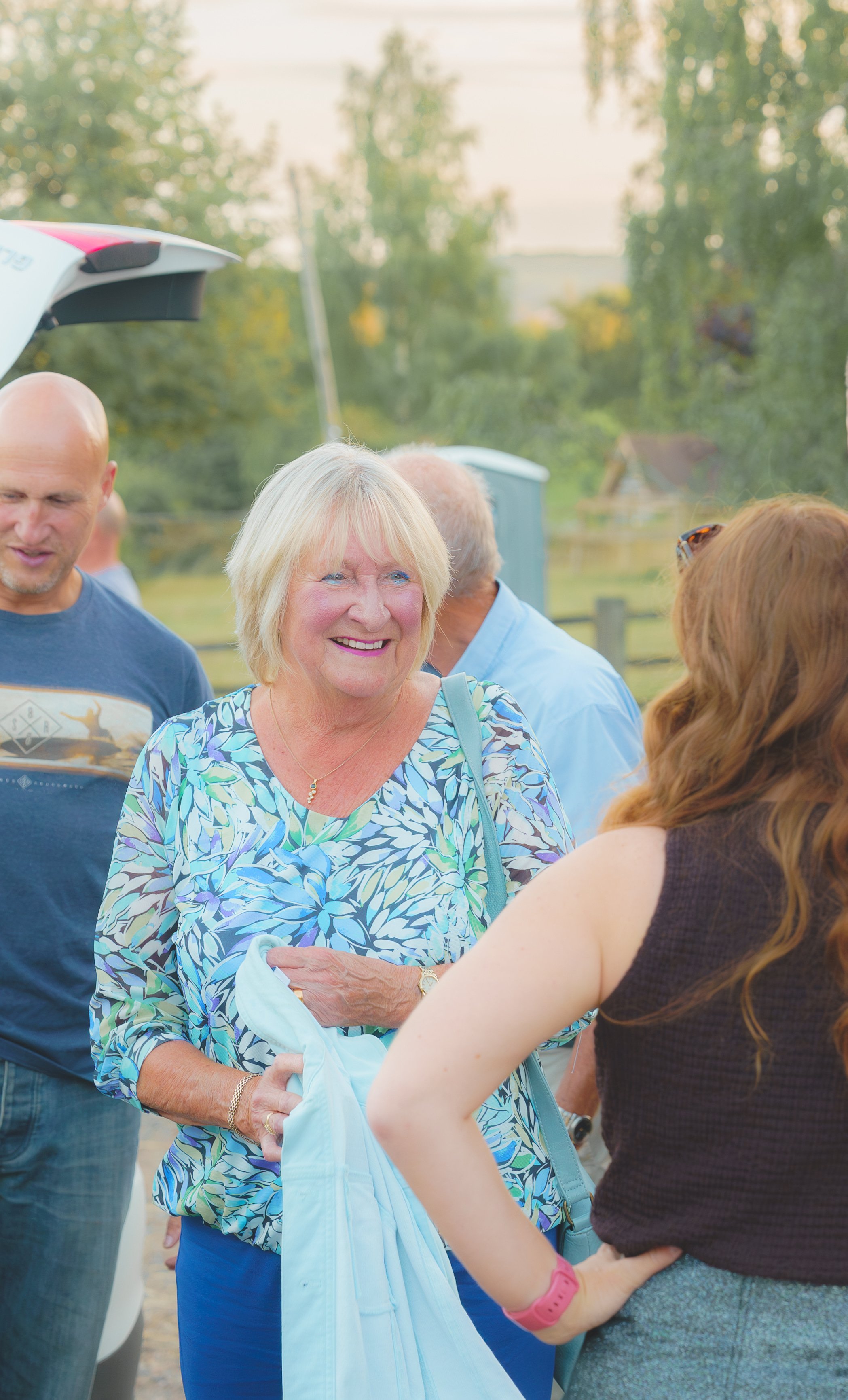 A group of people gathering outdoors, with a woman in a blue patterned blouse smiling and talking to a woman in a dark sleeveless top, holding a light-colored jacket in her hand. Others are visible in the background, with trees and a blue portable re