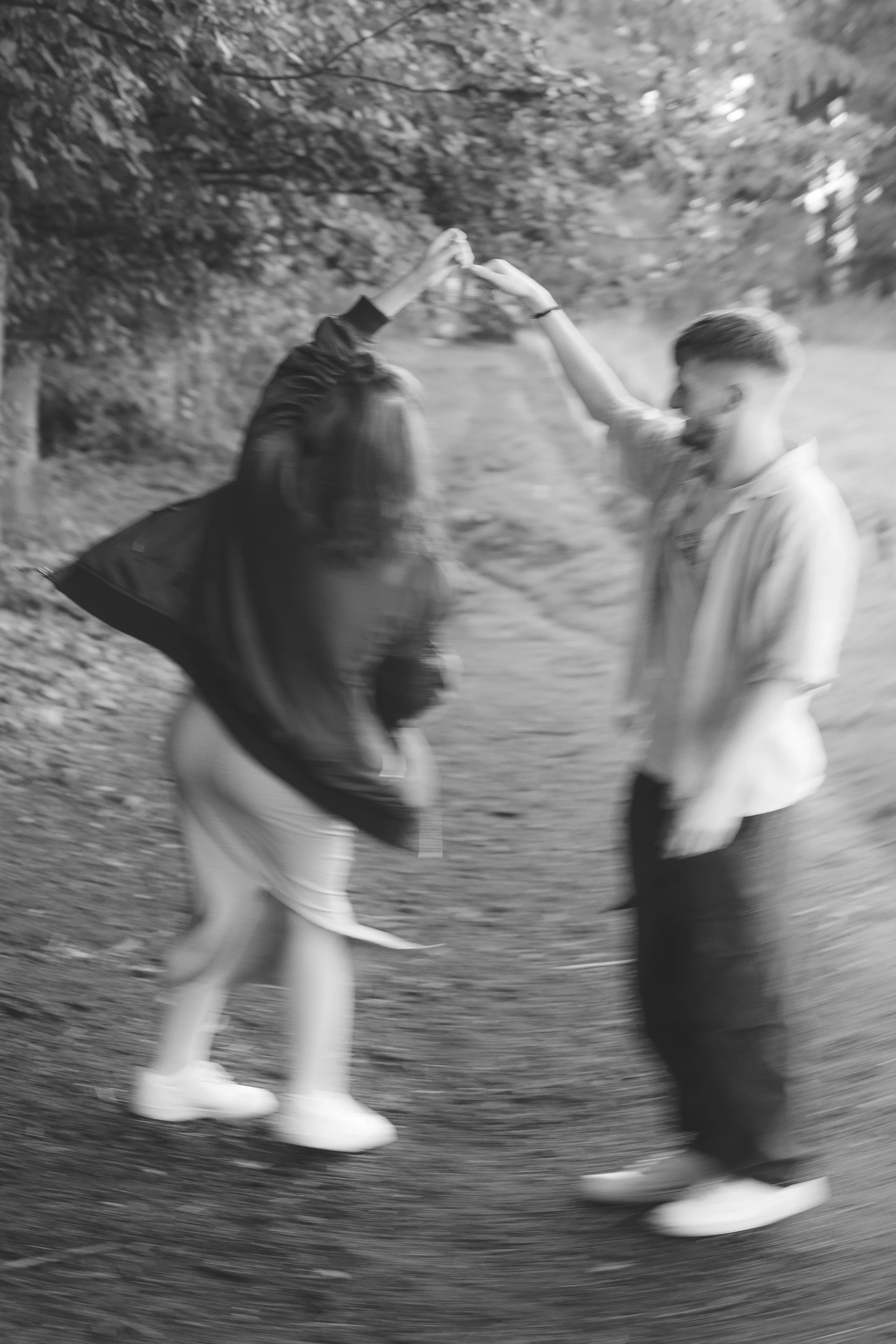 A blurry black and white photo of two people dancing outdoors with a tree and path in the background. They are holding hands, and their bodies are in motion.