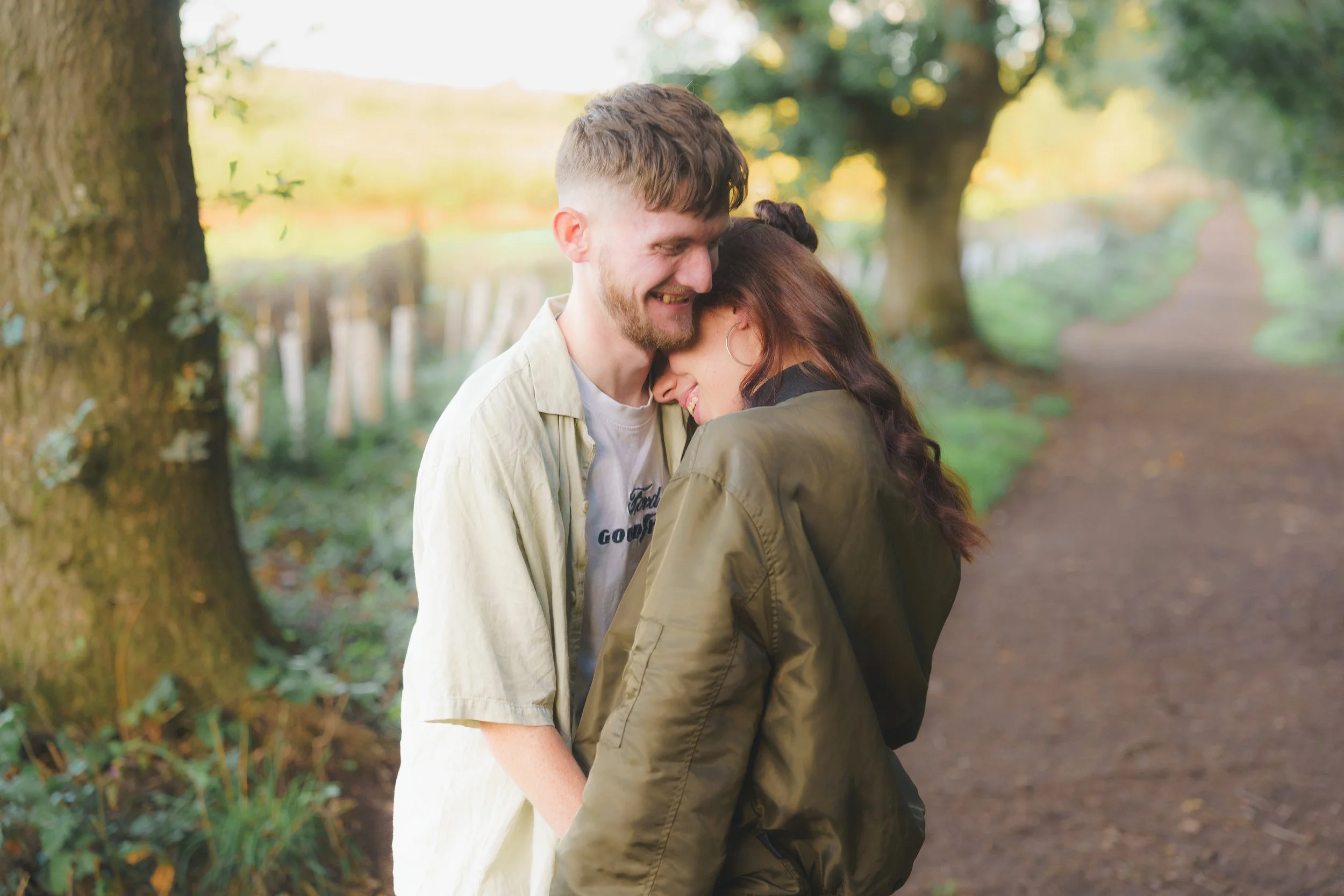 A young couple sharing a close, joyful moment outdoors on a nature trail surrounded by trees and greenery during daytime.