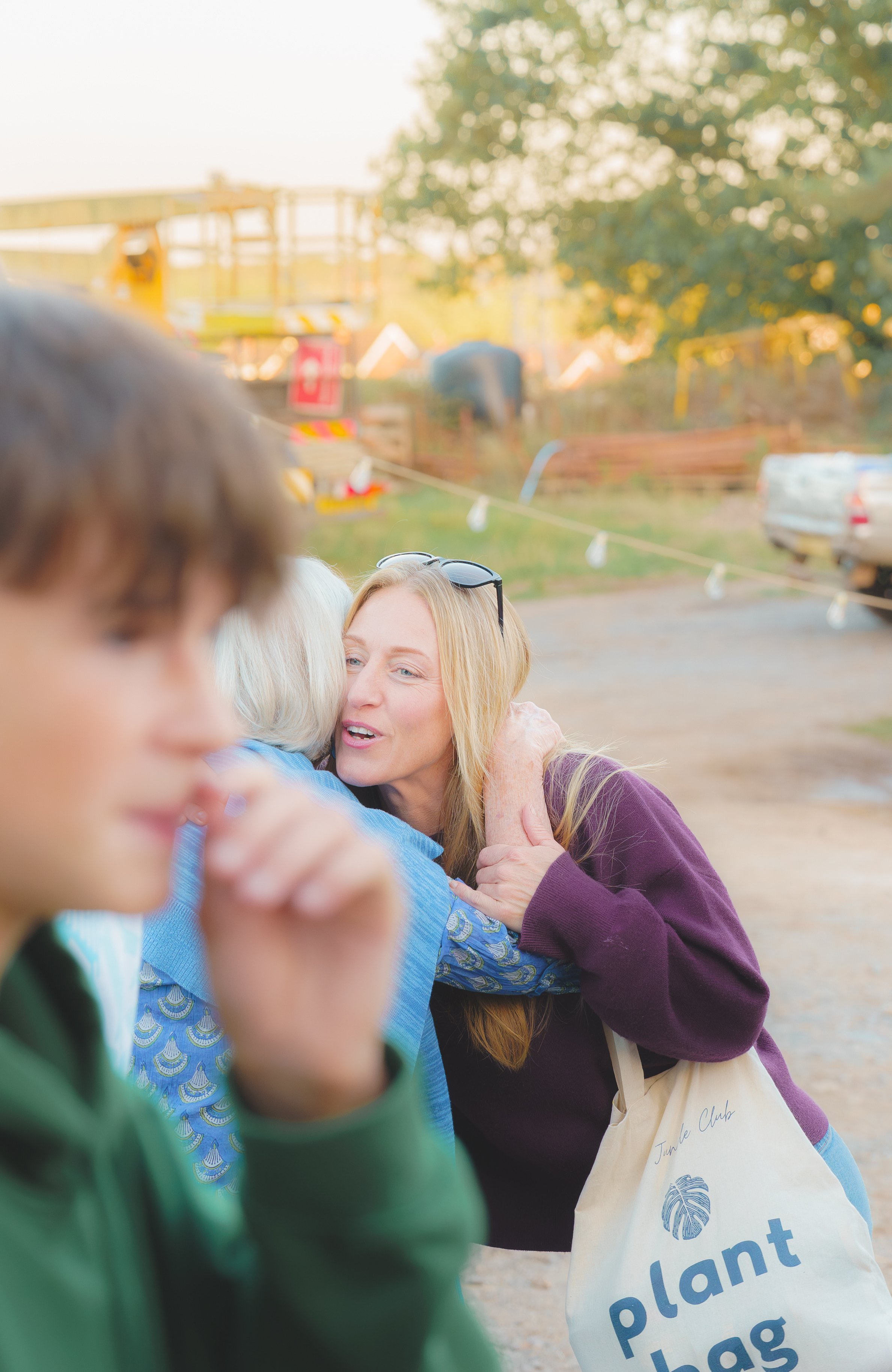 People hugging outdoors during a sunny day, with a woman in a purple sweater and sunglasses smiling at the center, and others partially visible in the foreground. In the background, there are trees, a parked car, and construction equipment.
