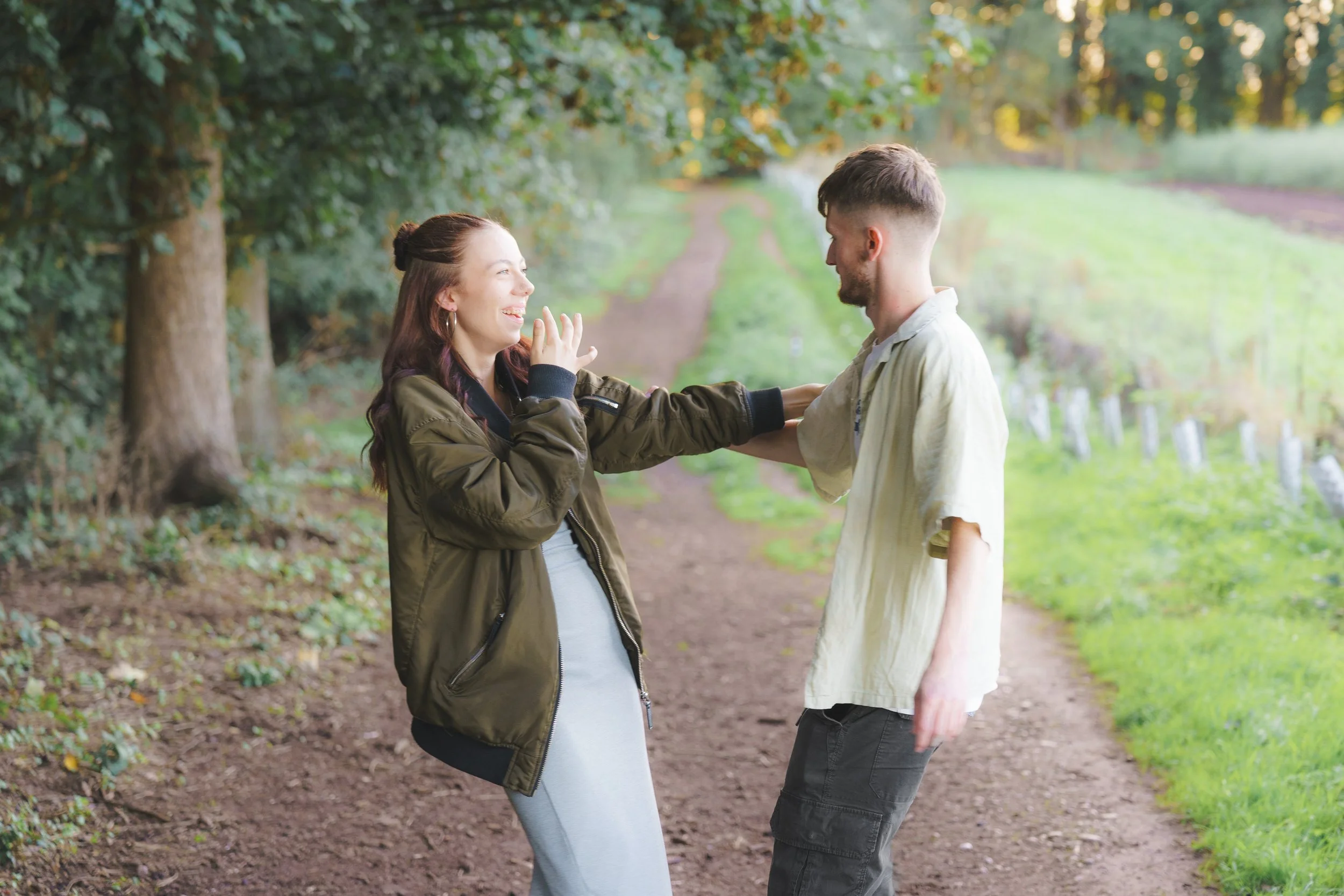 A young man and woman are outdoors in a park, standing on a dirt path amidst trees, smiling and appearing happy, with the woman wearing a green jacket and the man wearing a light-colored shirt.