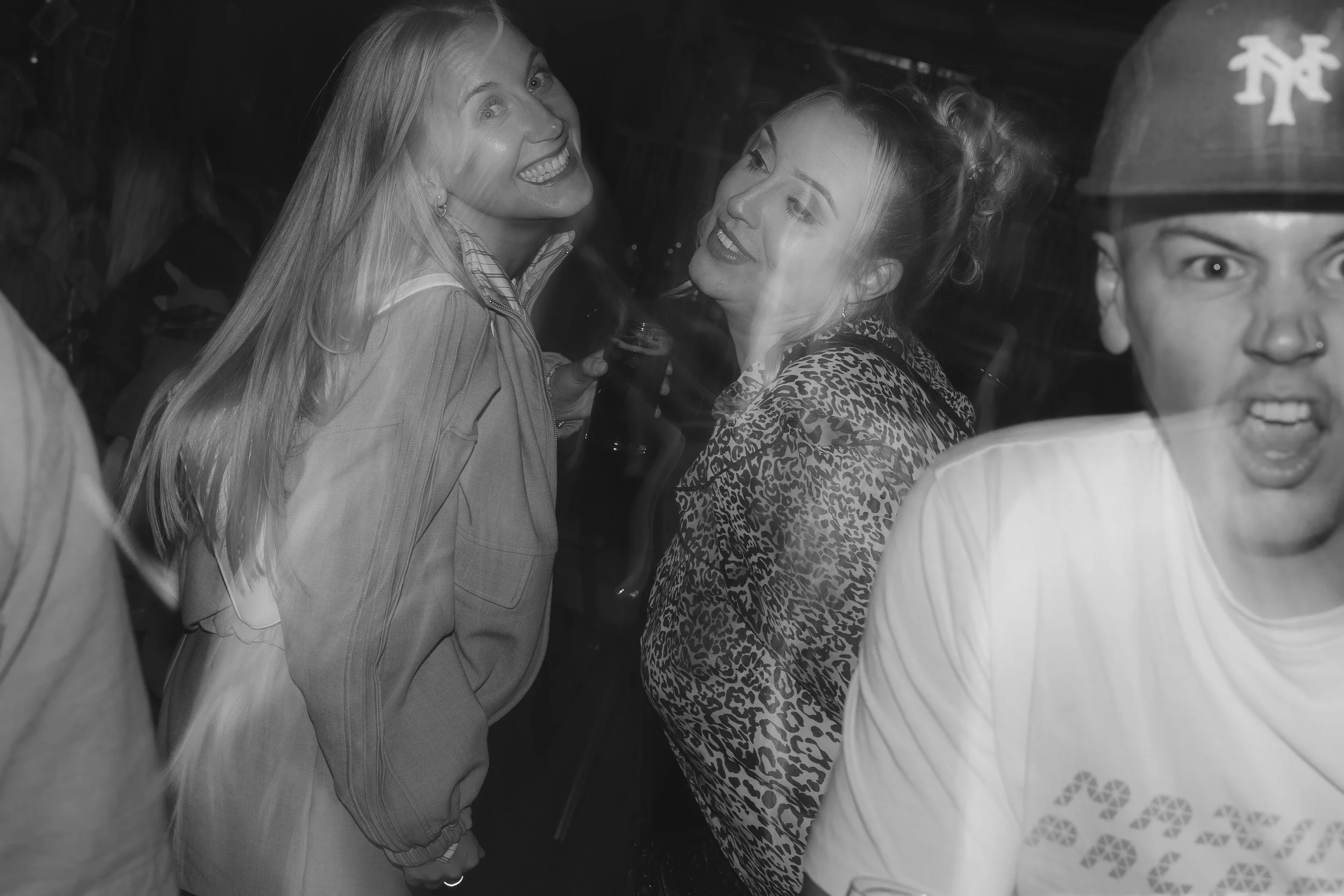 Three women smiling and talking at a social gathering, with one holding a drink, in a dimly lit setting.