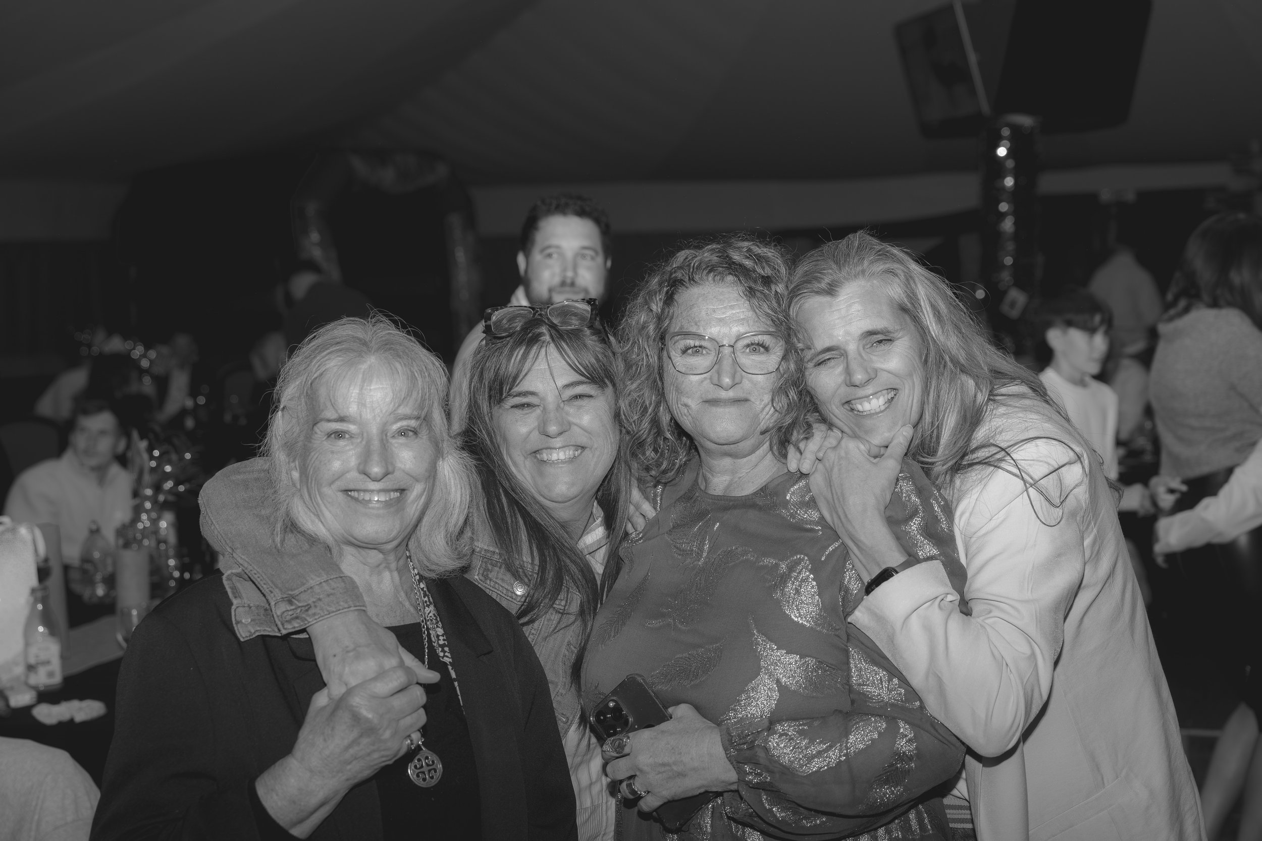 Four women smiling and hugging each other at a social gathering, with one woman holding a phone, in a room with other people in the background.