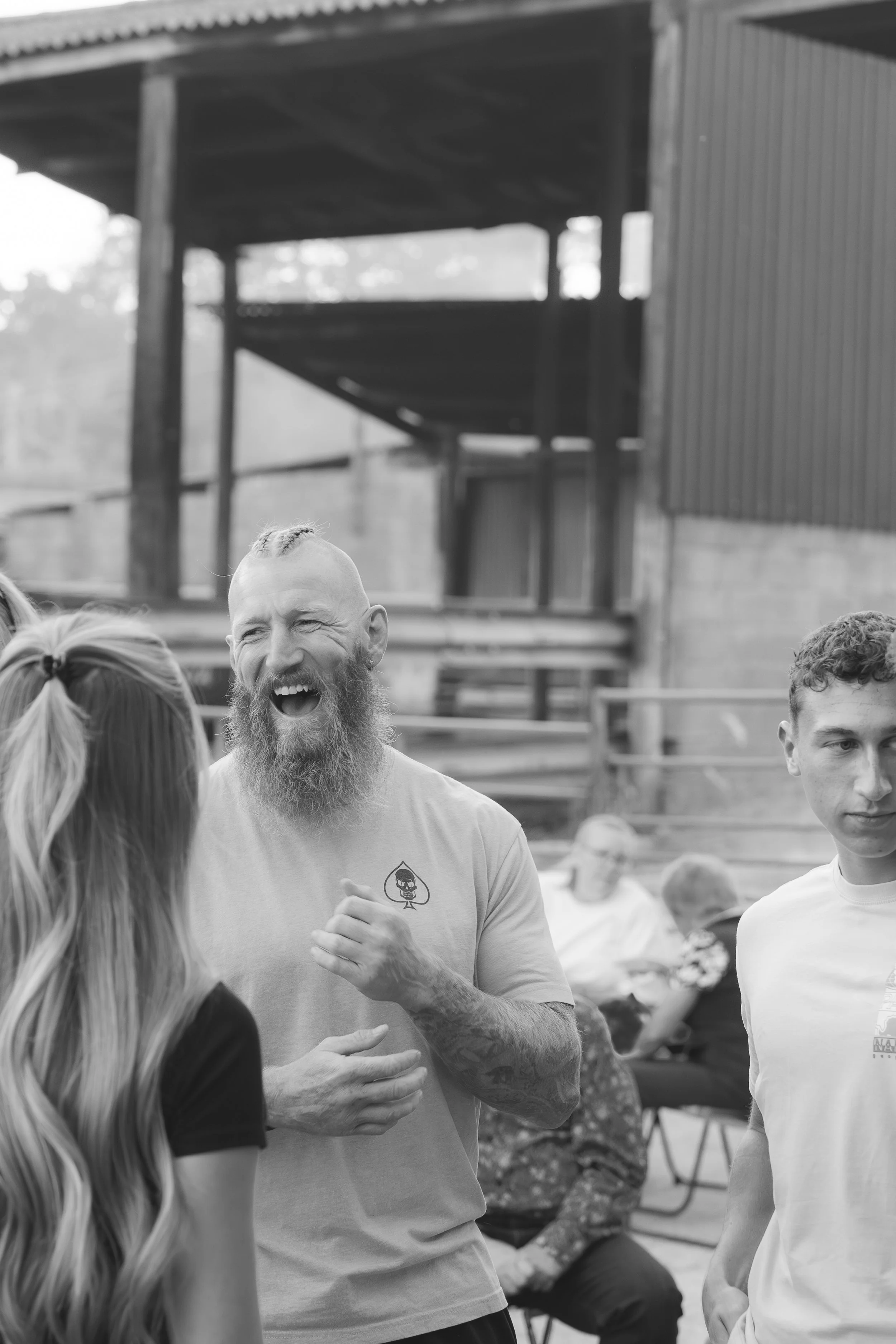 People gathered outdoors, smiling and engaging in conversation. One man in the center with a beard and short hair appears to be laughing.