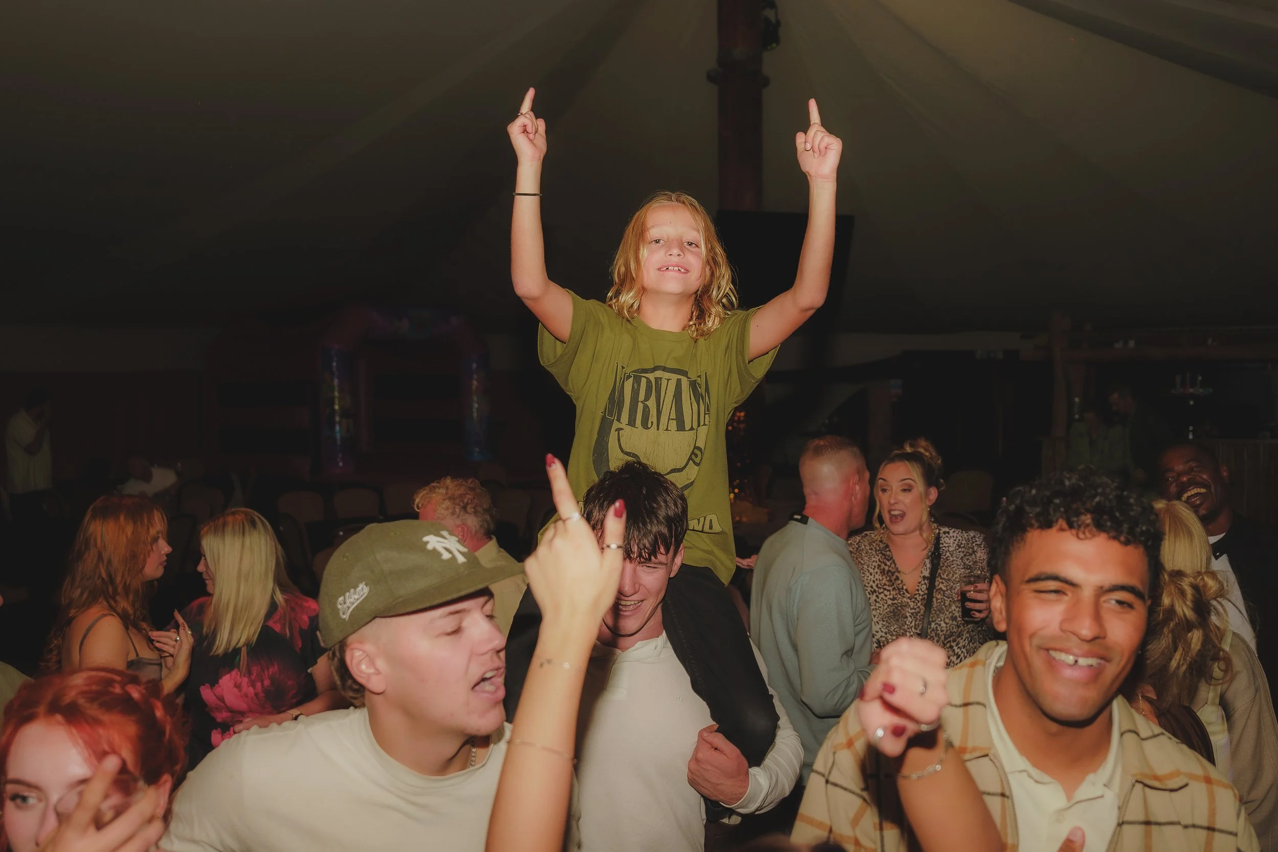 A group of people celebrating at a party, with a woman standing on someone's shoulders and raising her arms.