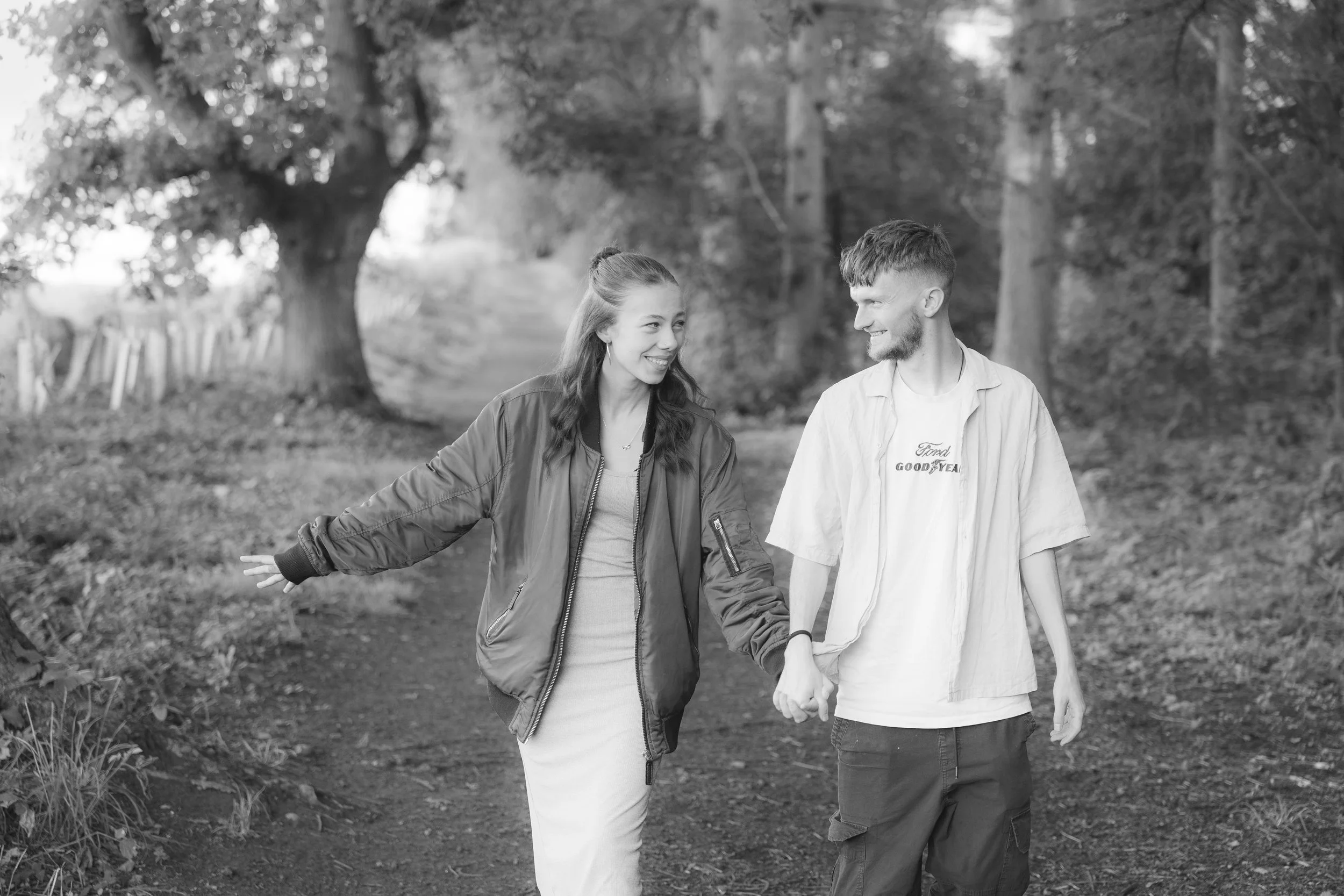 A black and white photo of a young couple walking outdoors hand in hand in a wooded area, smiling at each other.