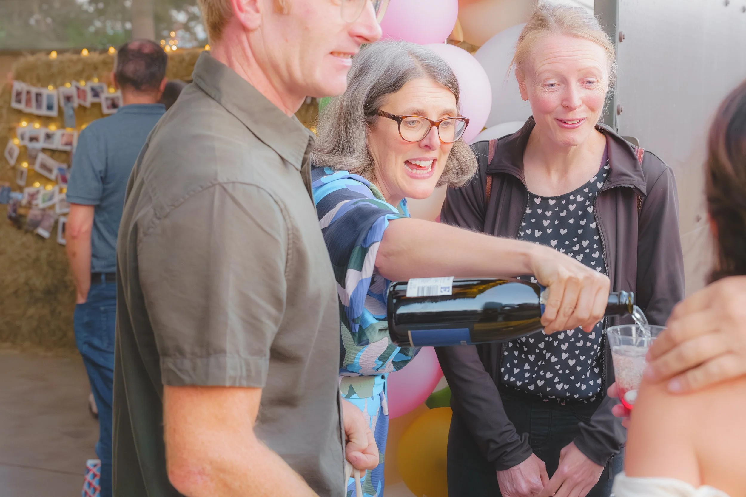 People socializing at a gathering with balloons and photo decorations in the background. One woman is pouring a drink from a wine bottle, smiling, while others are participating in the event.