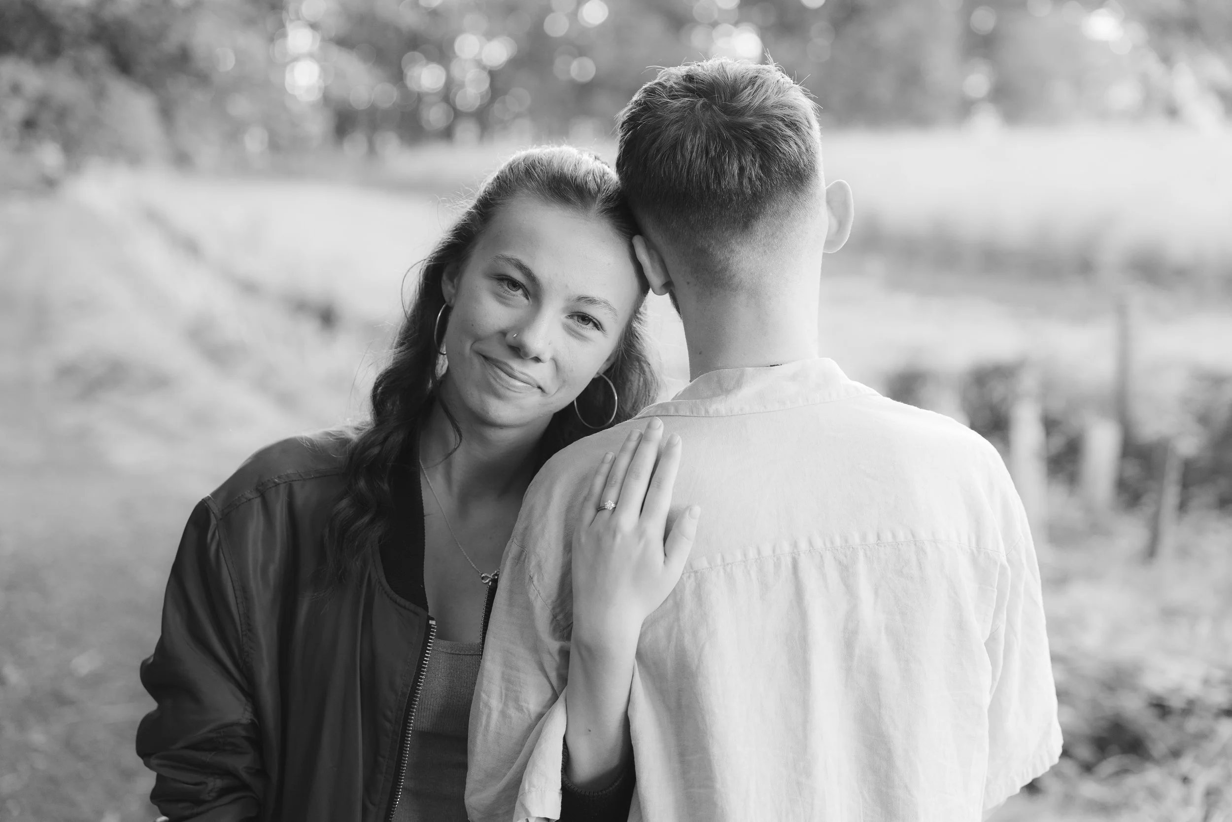 A black-and-white photo of a woman with long hair and earrings, embracing a man with short hair and a fade haircut outdoors. The woman is smiling gently, her hand resting on the man's shoulder, showing an engagement ring. The background is blurred with trees and natural scenery.