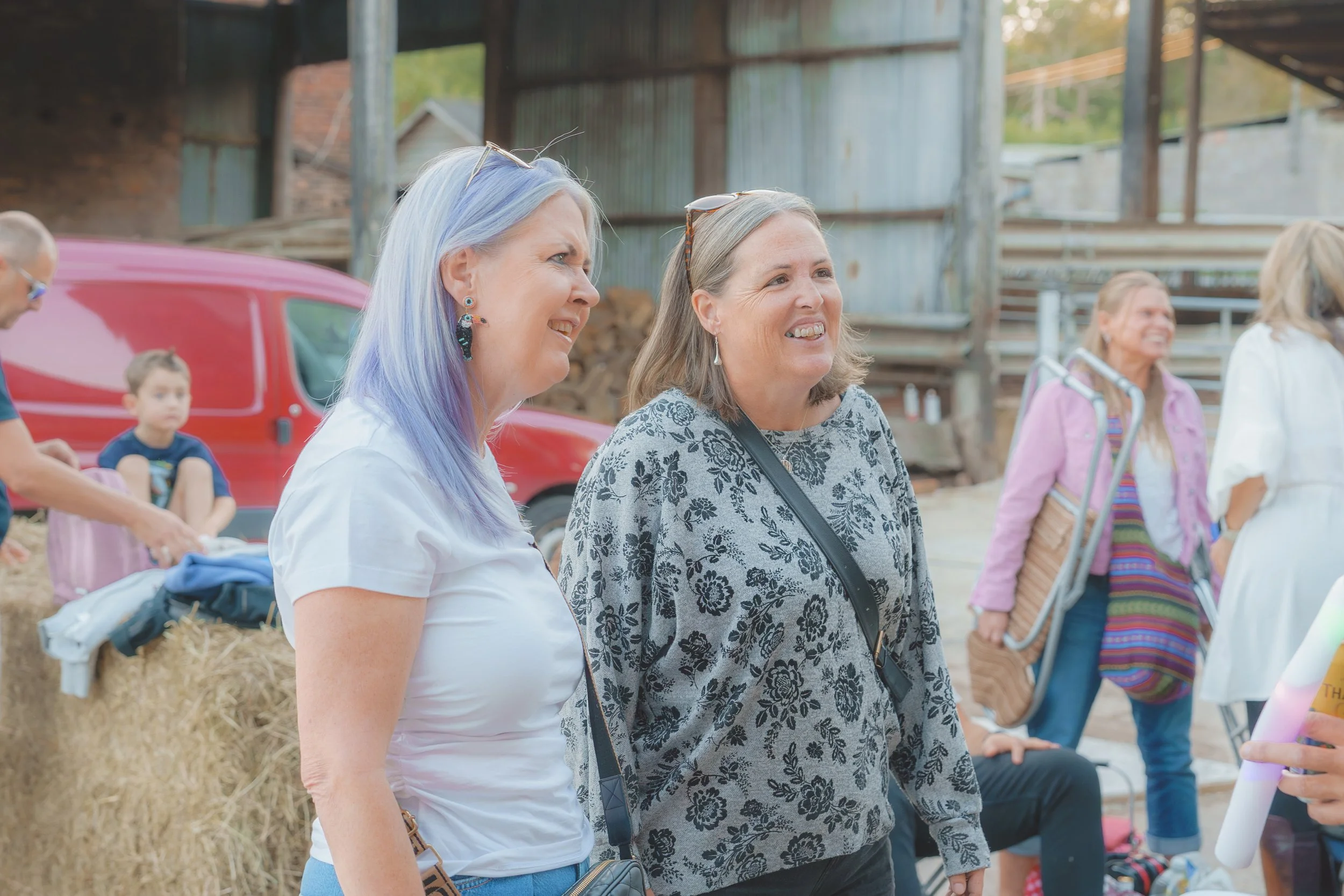 Two women smiling and standing near each other outdoors, with other people and a rustic barn in the background.