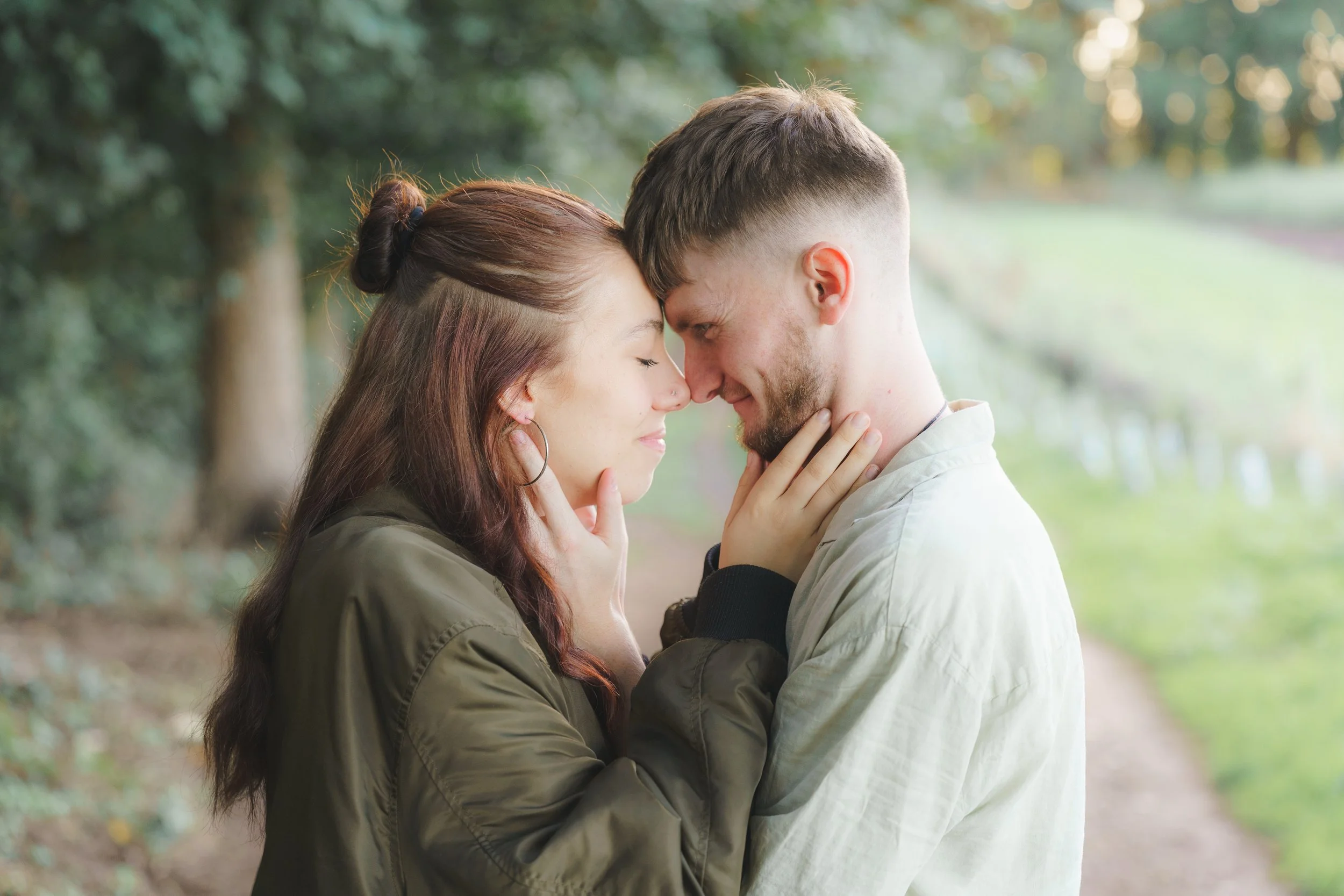 A couple with their foreheads touching, eyes closed, in a park with trees in the background.