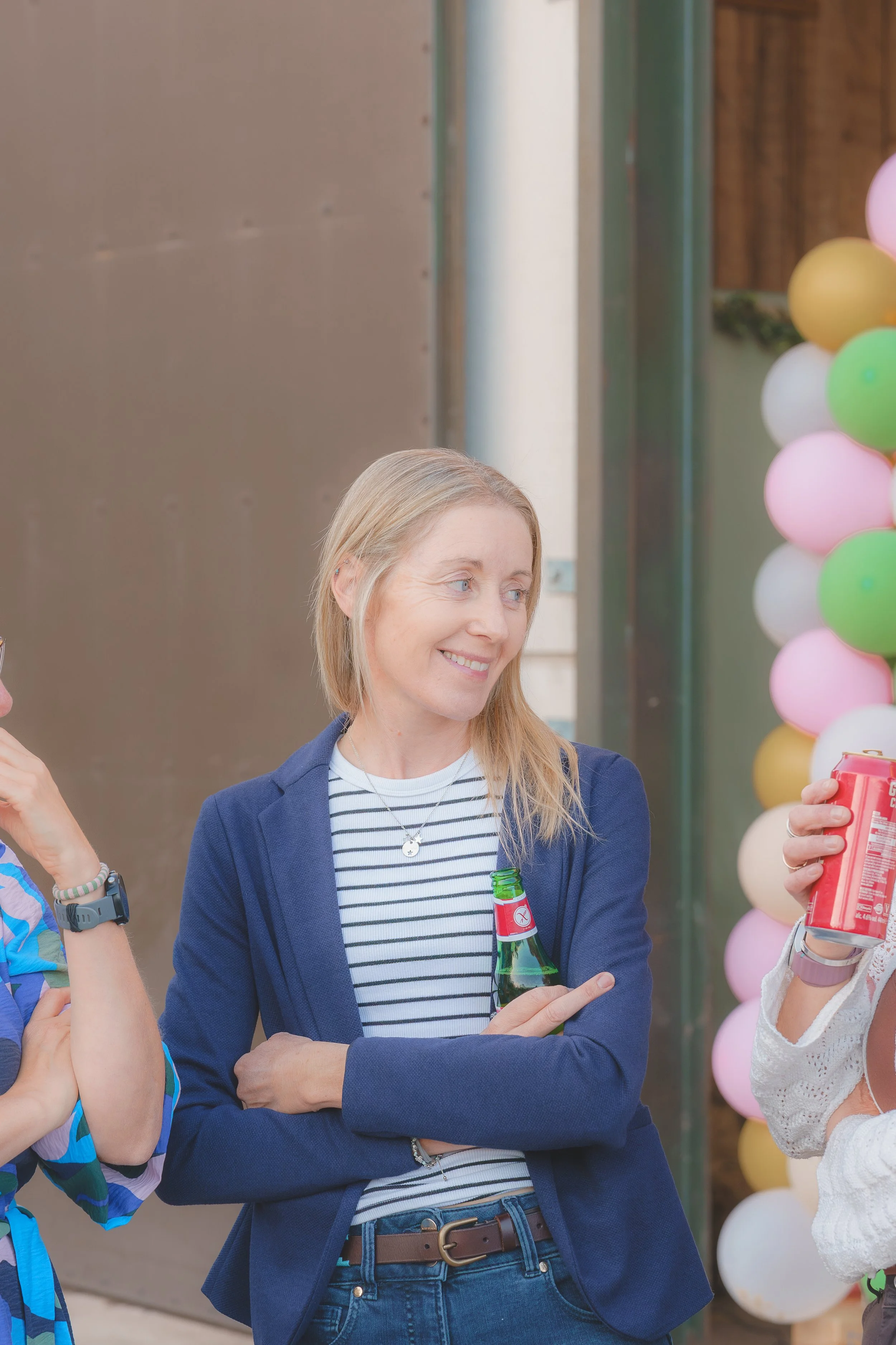 A woman with blonde hair wearing a navy blazer and striped shirt, holding a green bottle, smiling and talking to someone at an outdoor party decorated with pink, yellow, and white balloons.