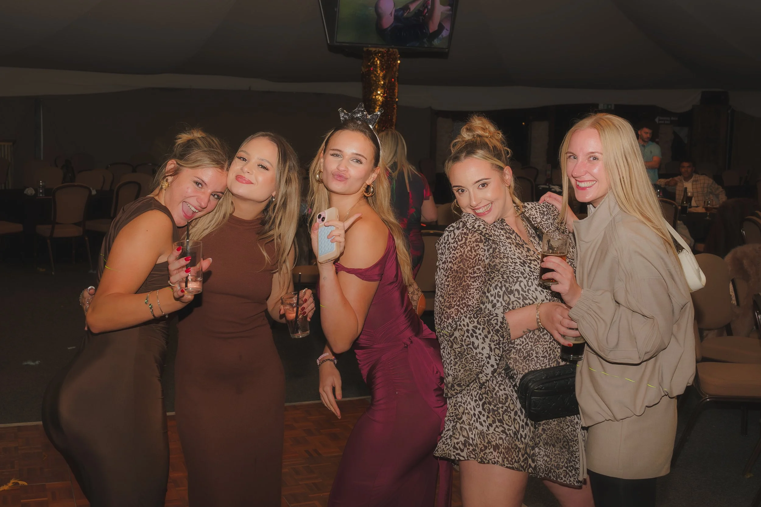 Group of six women smiling and posing for a photo at a celebration or party, holding drinks, with one woman wearing a headband with silver stars, in an indoor venue with chairs and tables in the background.