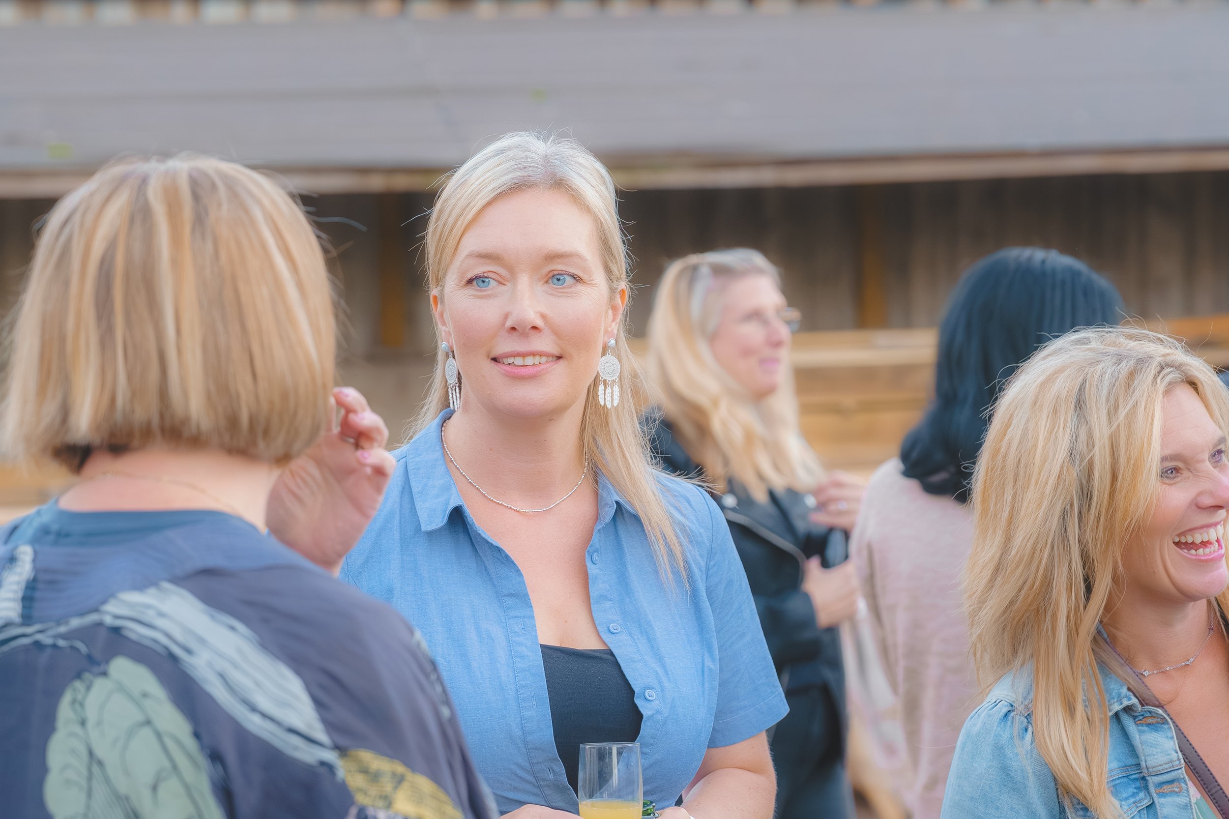 A group of women socializing outdoors, with the focus on a woman with blonde hair, blue eyes, wearing earrings and a blue shirt, engaging in conversations.