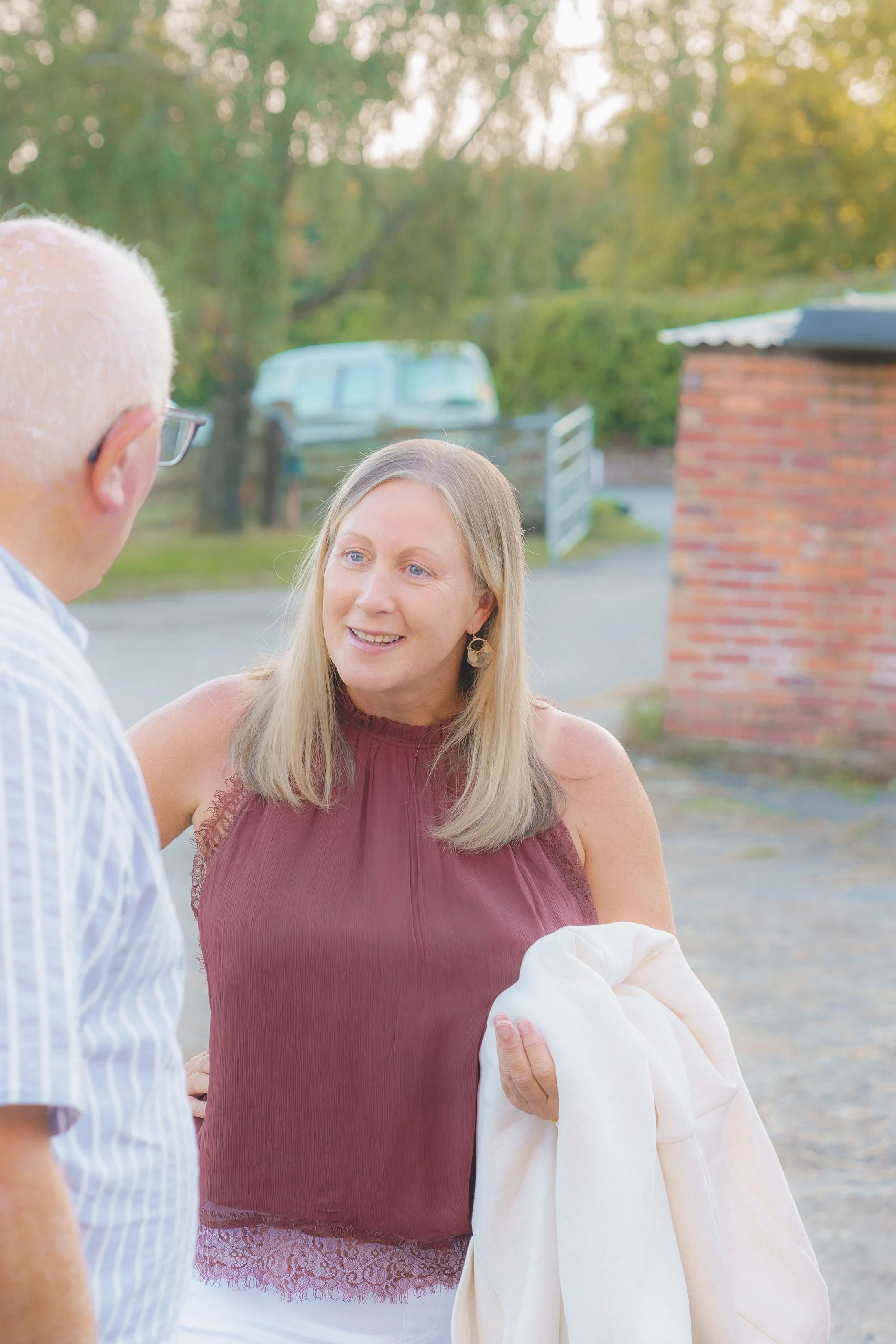 A woman holding a baby wrapped in a white blanket, standing outdoors near a brick structure during sunset, with a man in glasses and a striped shirt talking to her.