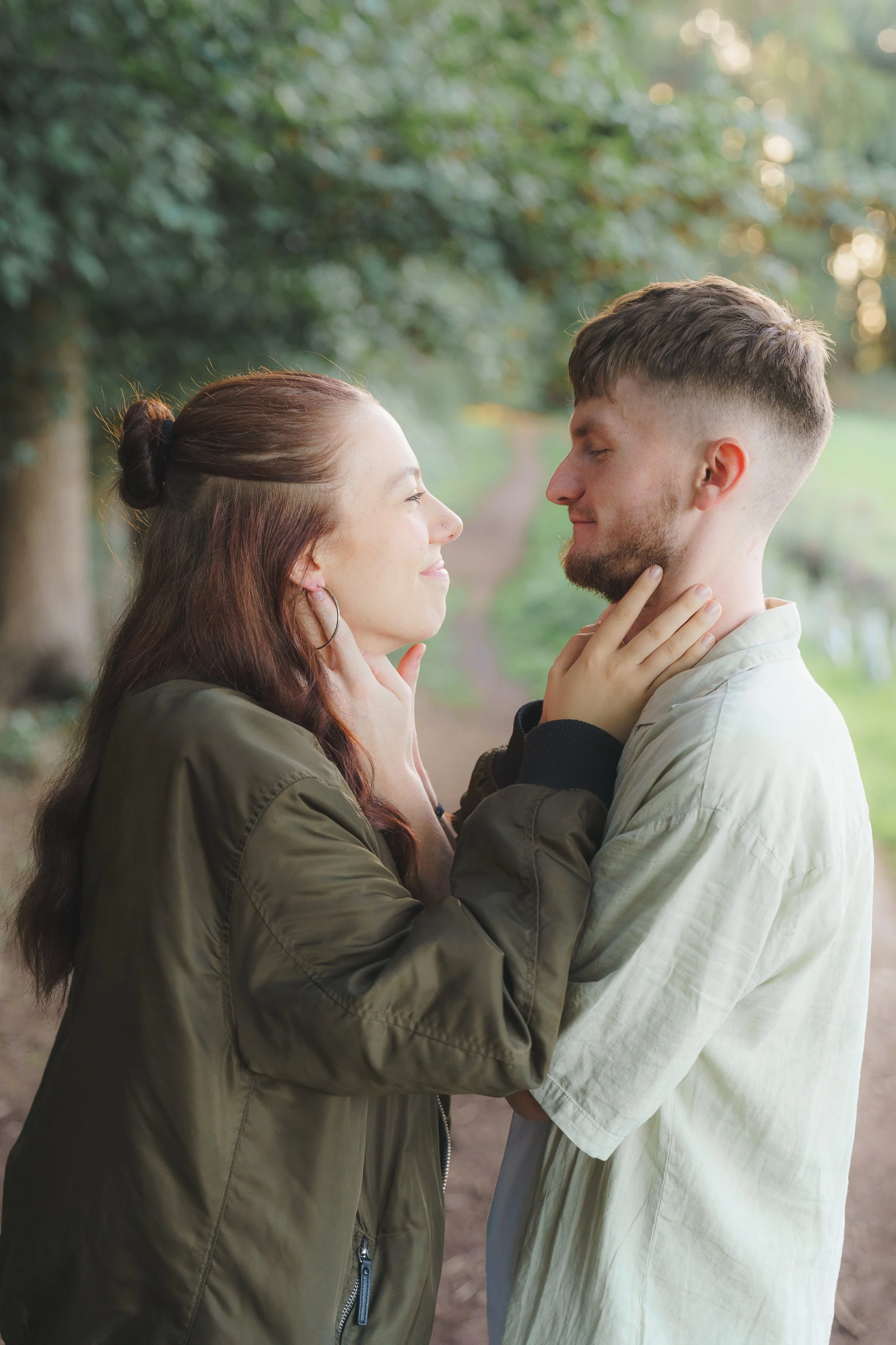 A couple standing close together outdoors with the woman touching the man's chin and the man holding her face, both with eyes closed, appearing happy and affectionate.