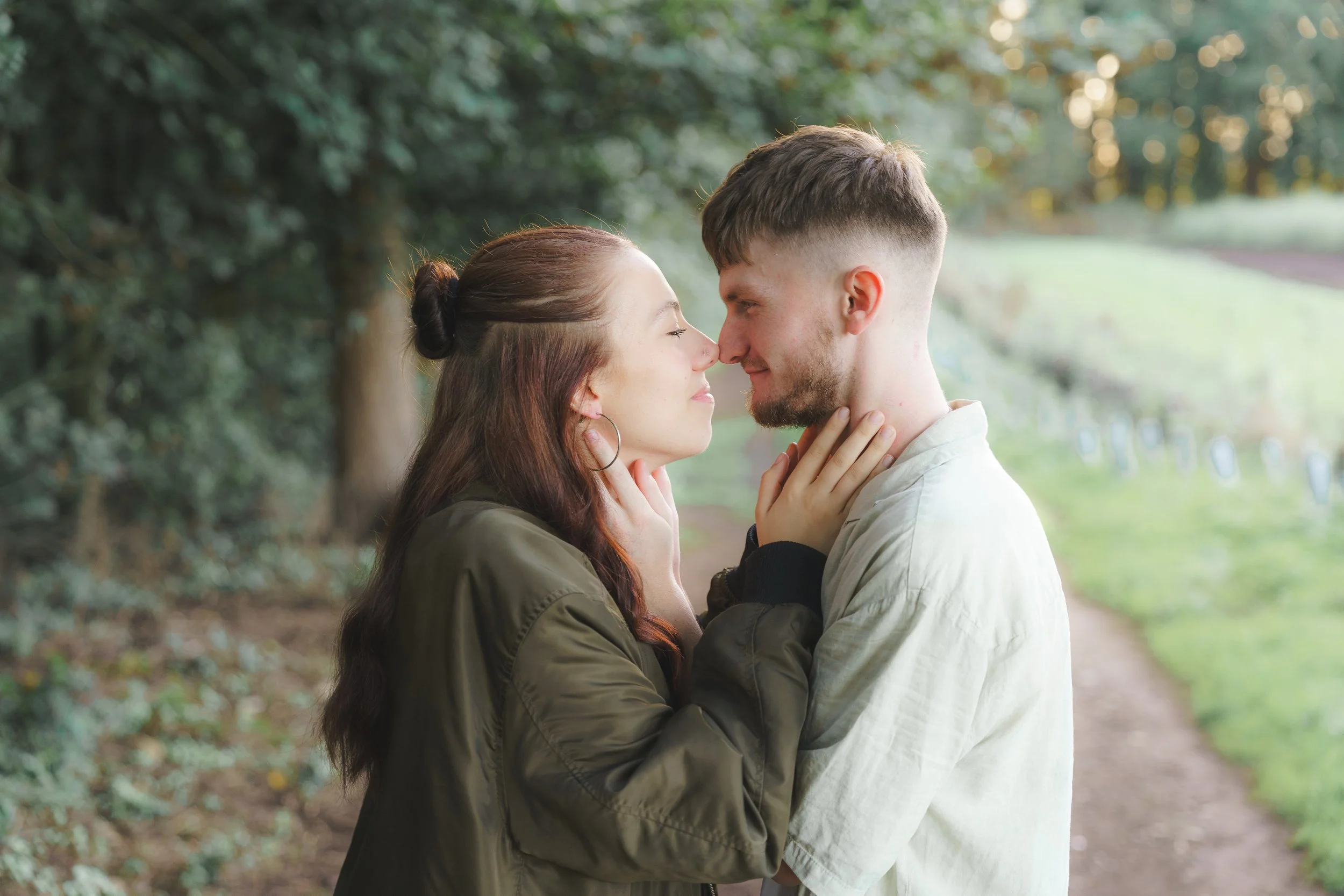A couple standing close together in a park, touching foreheads and holding each other's faces, surrounded by trees and greenery.