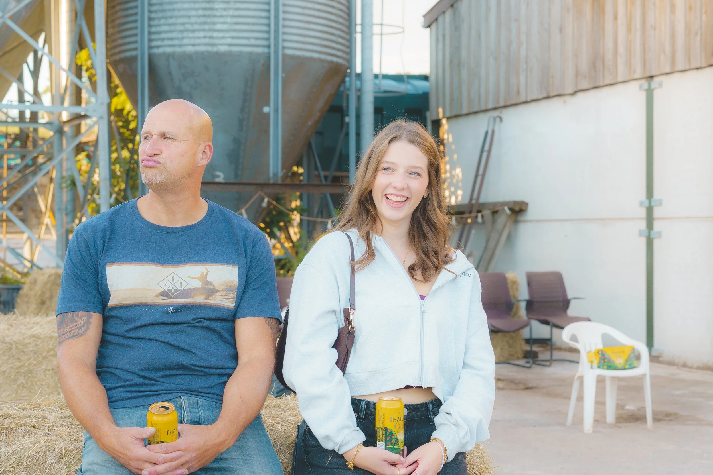 A man and woman standing outdoors, holding yellow cans, with farm or barn equipment in the background.