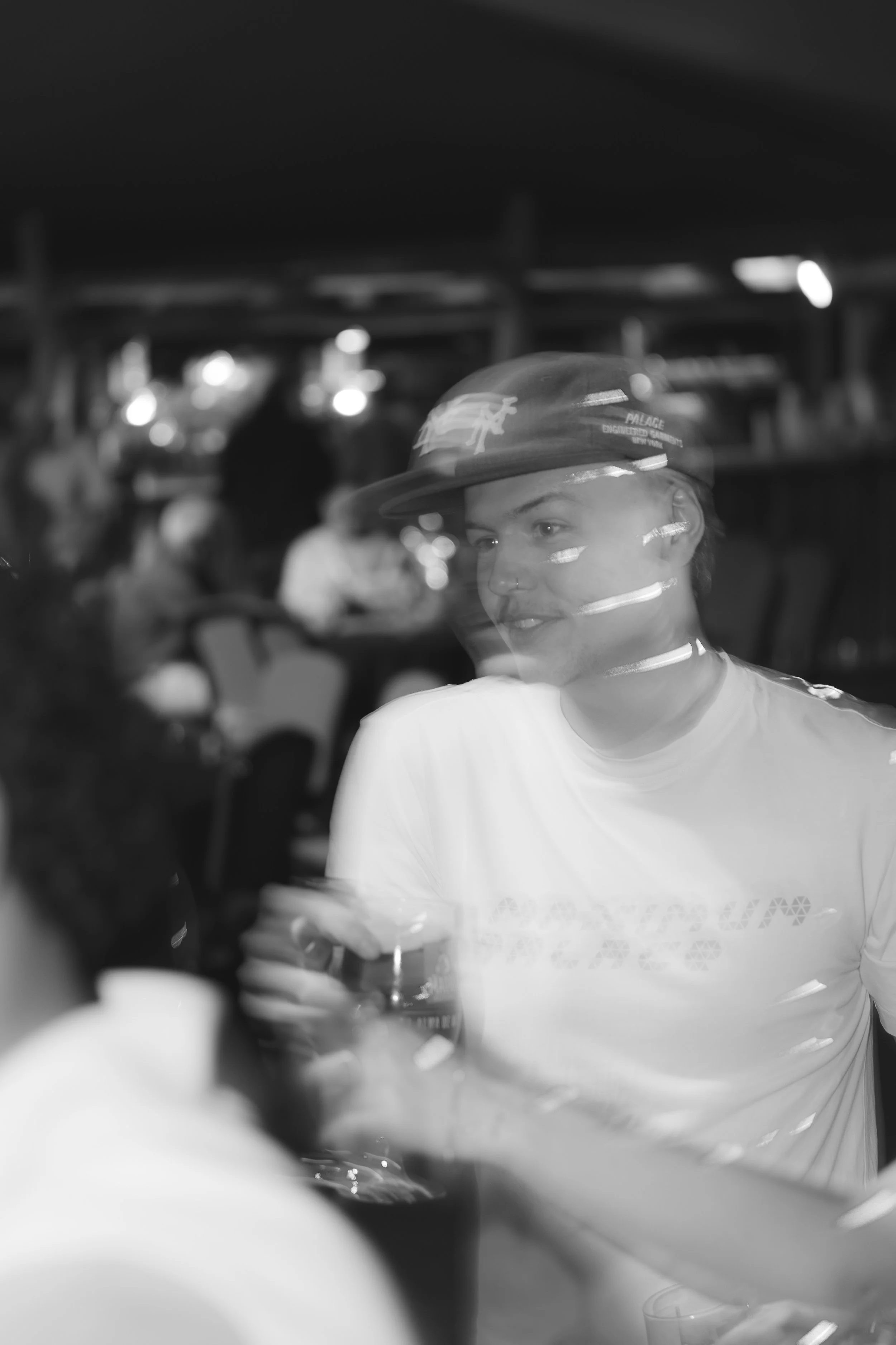 A black and white photo of a young woman wearing a hat and white t-shirt, sitting at a table holding a glass, with a blurred background of people and lights at night.