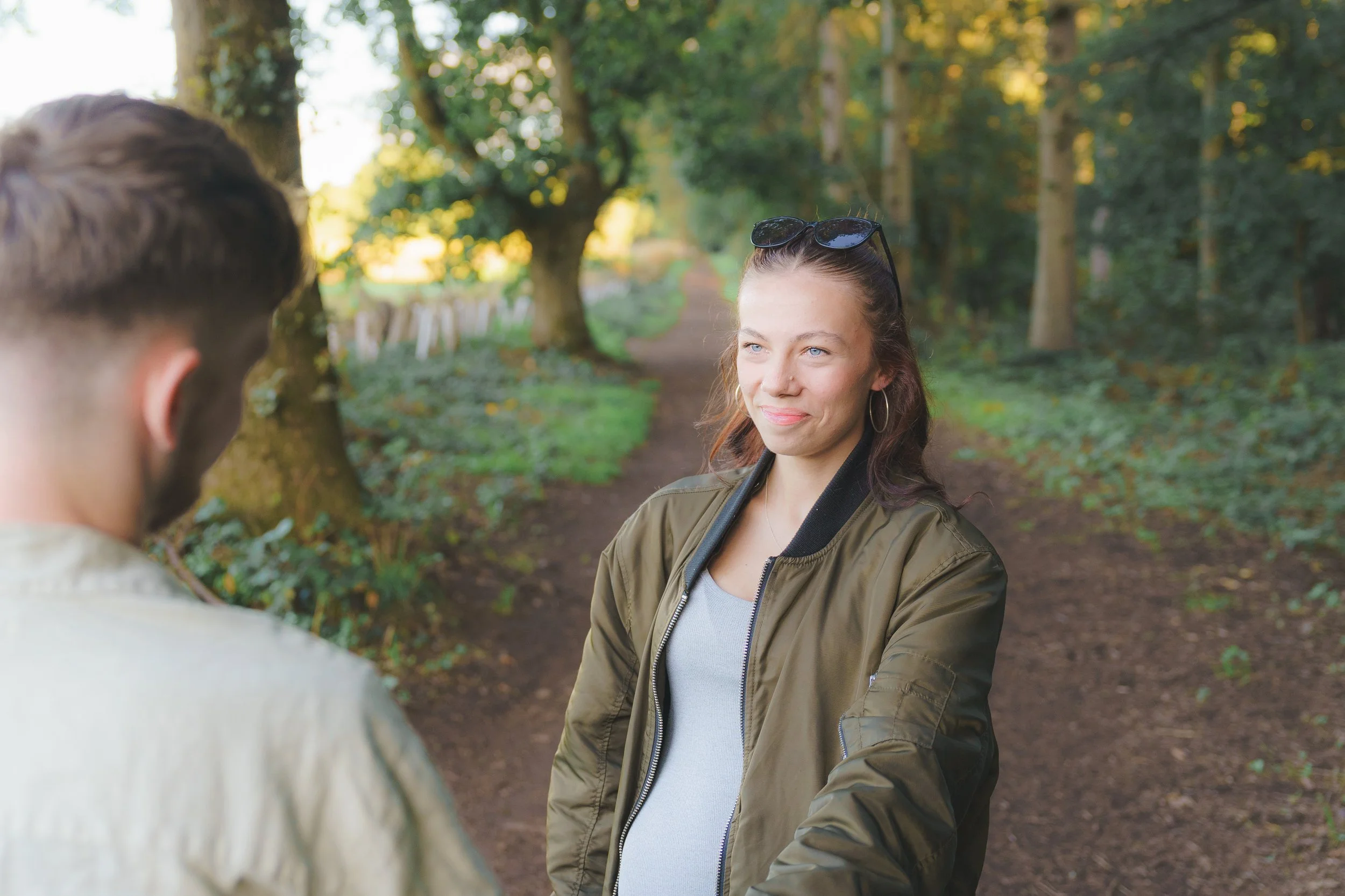 A young woman with blue eyes and brown hair tied back, wearing sunglasses on her head, a green jacket, and hoop earrings, is smiling and looking at a man with brown hair, in an outdoor park setting with trees and a dirt path.