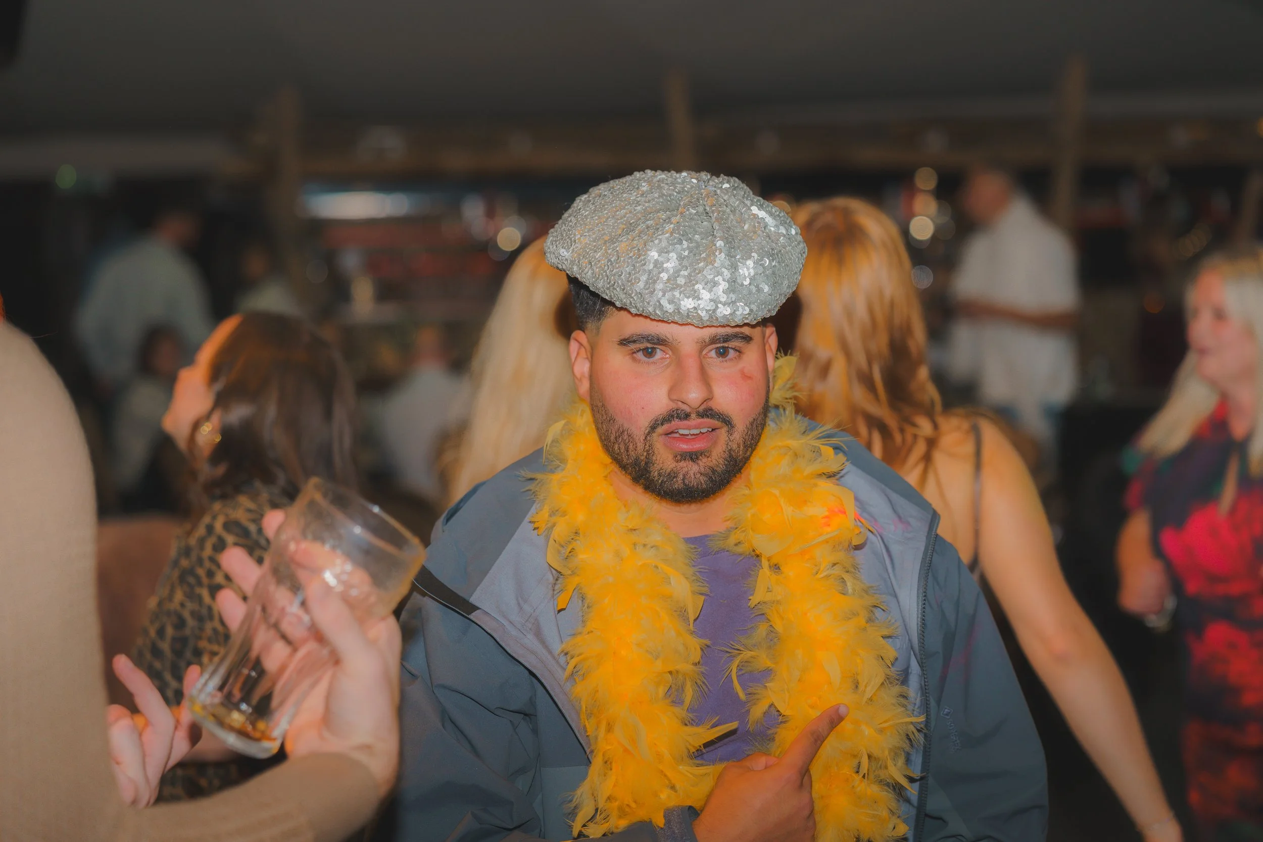 A man with a beard and blue eyes wearing a shiny silver sequined beret and a yellow feather boa at a party, surrounded by people.