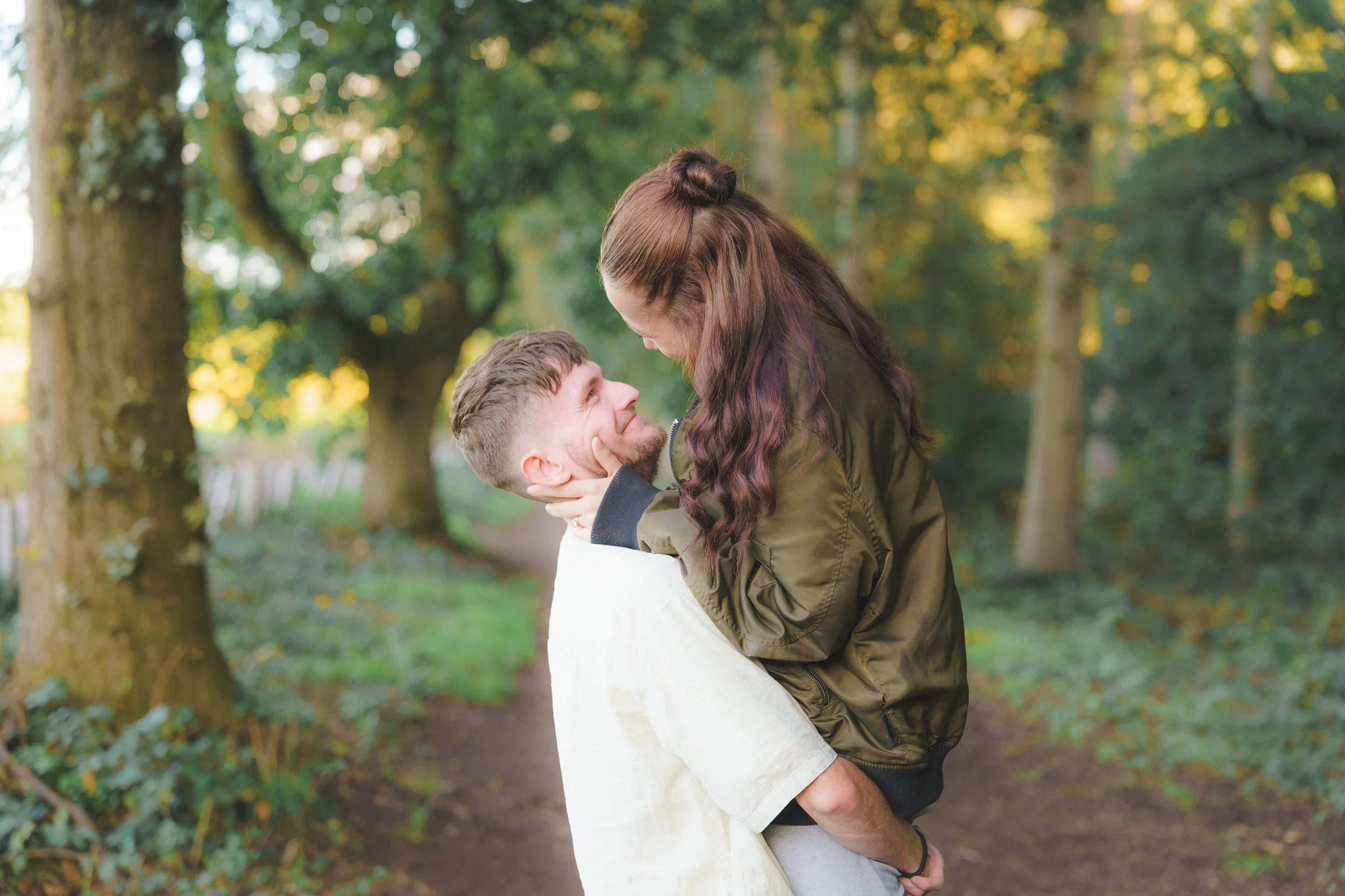 A couple in a park, the man lifting the woman, both smiling, surrounded by trees with green and yellow leaves.