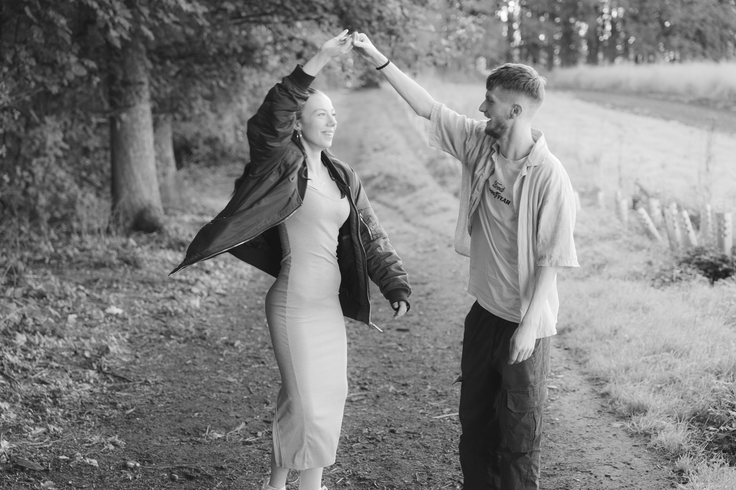 A young man and woman dancing and holding hands in an outdoor park with trees in the background, captured in black and white.
